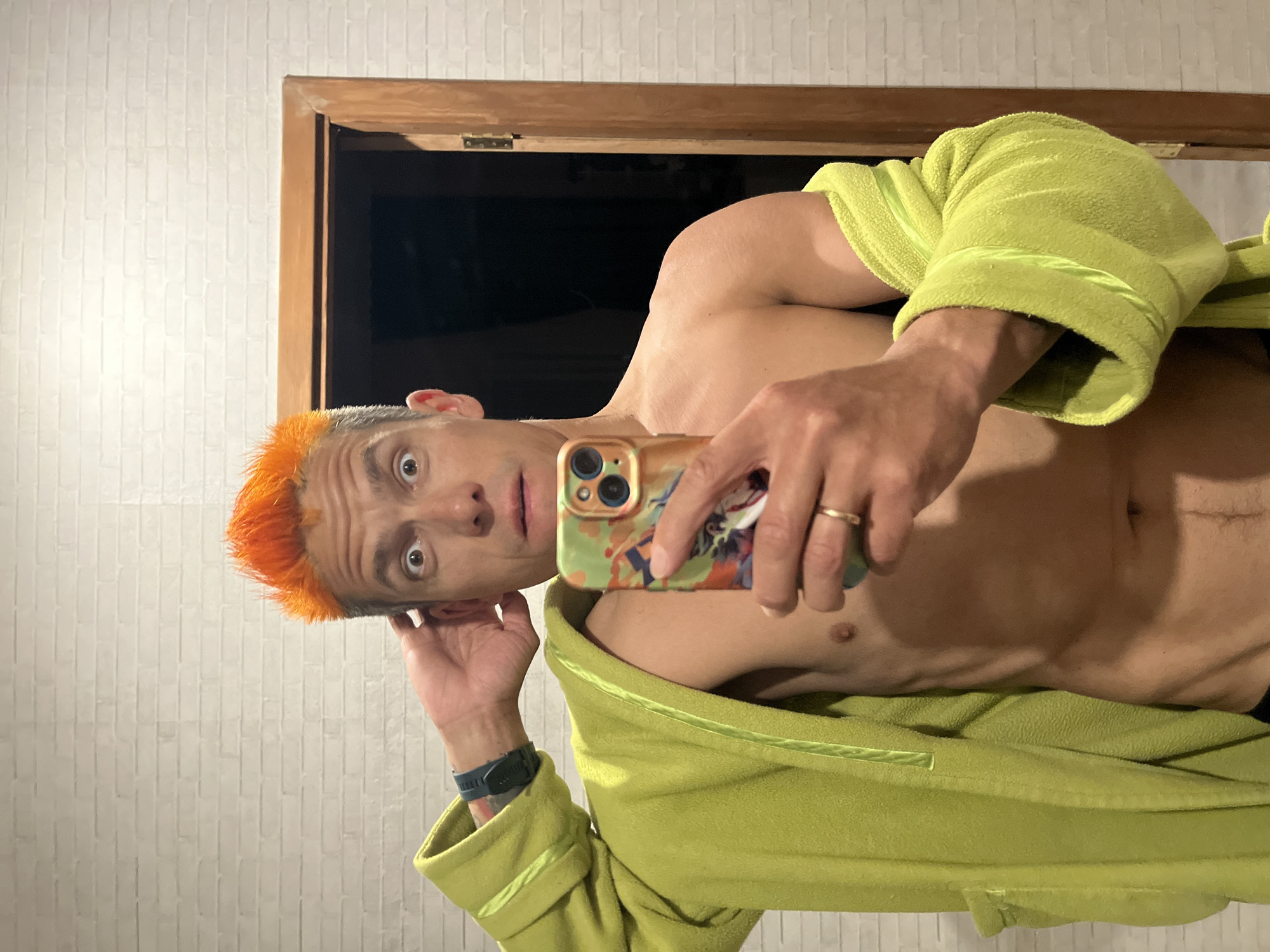 A shirtless man with orange hair takes a selfie in a bathroom. He is wearing a lime green robe and has a watch on his left wrist. He holds an iPhone with a patterned case. The background includes a white brick wall and a wooden-framed mirror reflecting a dark space. The location is identified as the city of Chaikovsky, Russia.