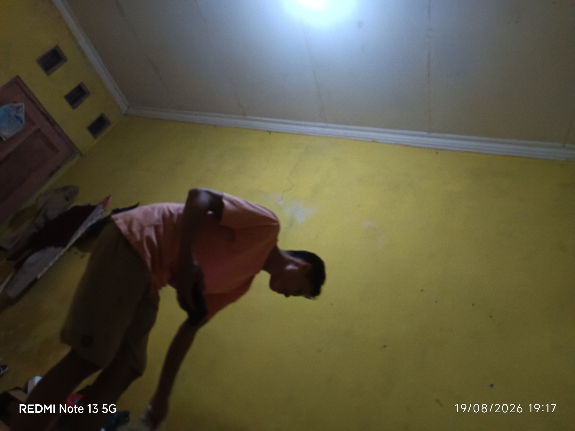 The image captures a low-angle shot of a young male with dark hair and skin, wearing a salmon-colored t-shirt and light brown shorts. He is in a crouched or bent-over position, facing away from the camera and slightly to the right. His left hand is holding a dark object, possibly a phone, near his torso. The background consists of a yellow-painted wall, a white ceiling with a light fixture, and a wooden door on the left side. The floor is also visible. A timestamp in the bottom right corner reads "19/08/2026 19:17", and "REDMI Note 13 5G" is in the bottom left. The scene appears to be an indoor residential setting, likely a room in Talaga Sari, Indonesia.