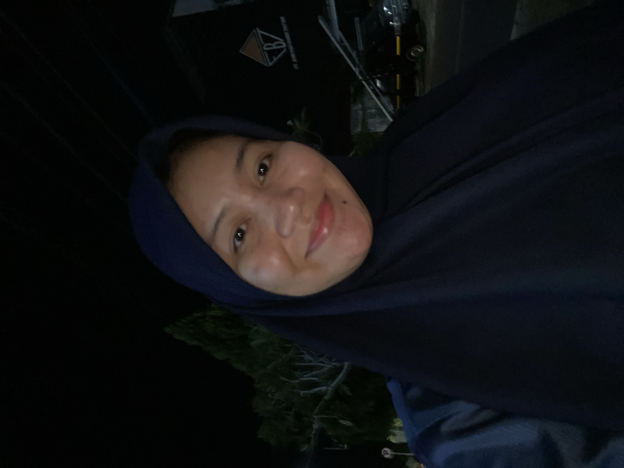 A woman wearing a navy blue hijab and a matching long-sleeved top is taking a selfie at night. She is smiling and looking at the camera. In the background, there is a tree, a building with a logo that reads "PT. ENERGI HIJAU TAPIN", and a black pickup truck. The scene appears to be outdoors, possibly in an industrial area or a parking lot, given the presence of the building and the truck. The lighting suggests it is dark, with artificial lights illuminating the subjects and the background. The location is identified as Banjarbaru, Indonesia.