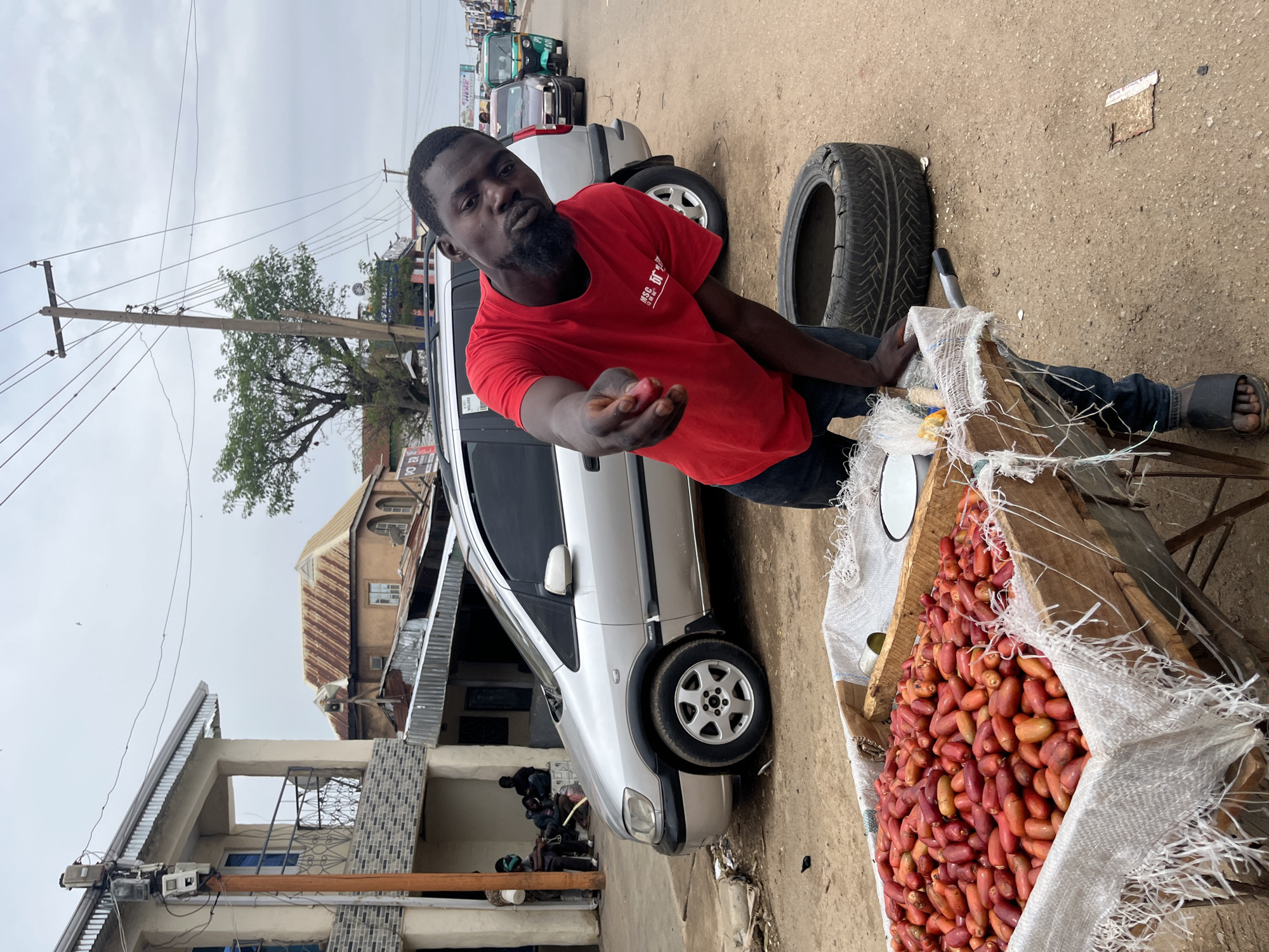 A man in a red t-shirt and jeans stands behind a wooden cart filled with red fruits, likely dates or a similar fruit, at a street market in Jos, Nigeria. He points a finger towards the camera while looking at the viewer with a direct gaze. A discarded tire lies on the ground behind him. In the background, a silver minivan is parked, and other vehicles, including a tuk-tuk, are visible on the street. Buildings with corrugated metal roofs and balconies line the street. The sky is overcast. The scene appears to be a typical street vendor setting in a populated urban area.