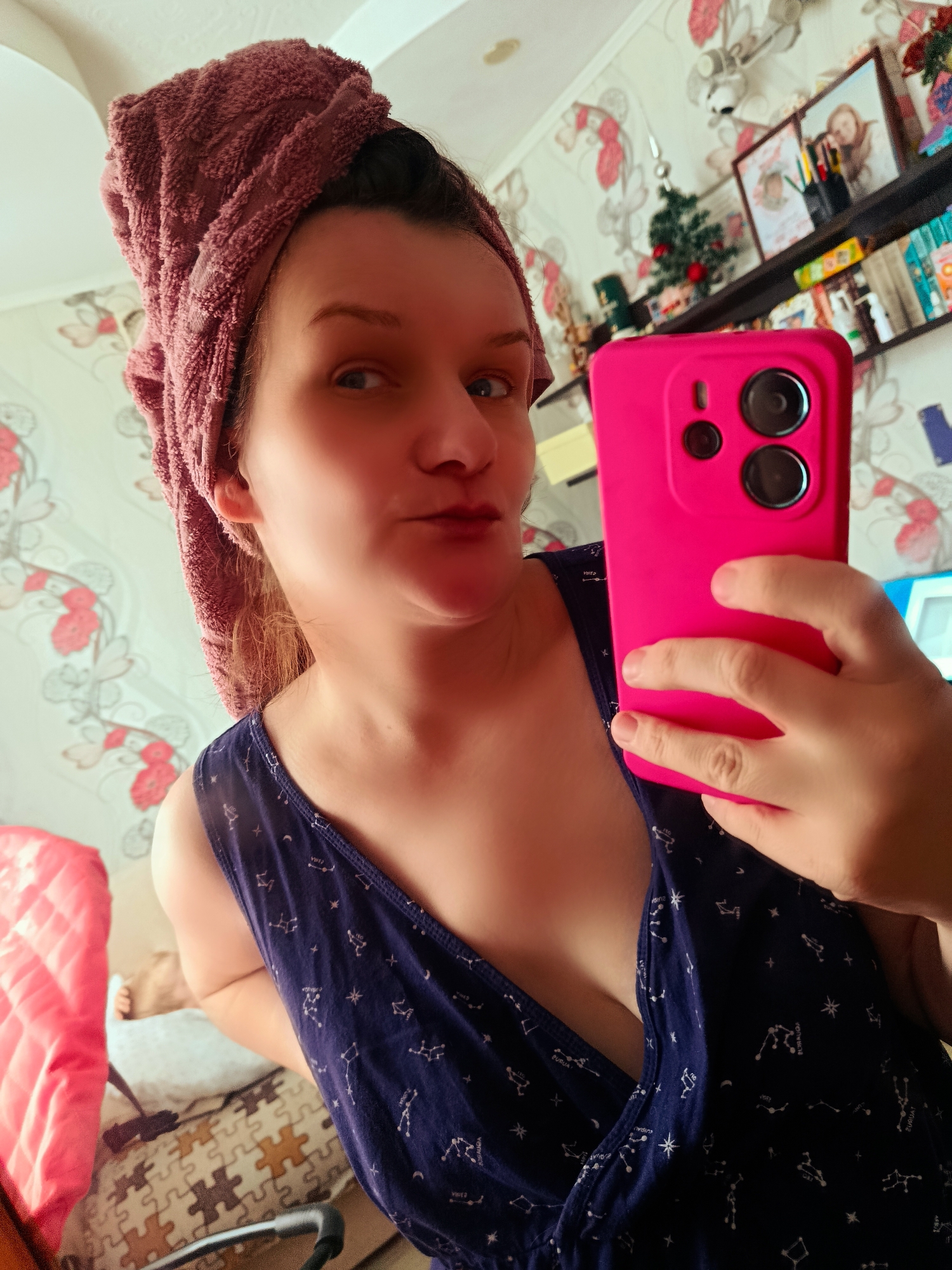 A woman is taking a selfie indoors. She has a pink towel wrapped around her head and is holding a pink smartphone in her right hand. She is wearing a blue top with constellation patterns. The background includes a patterned wallpaper, a shelf with various items, and a glimpse of a chair. There is no indication of this being in Фоки, Russia.
