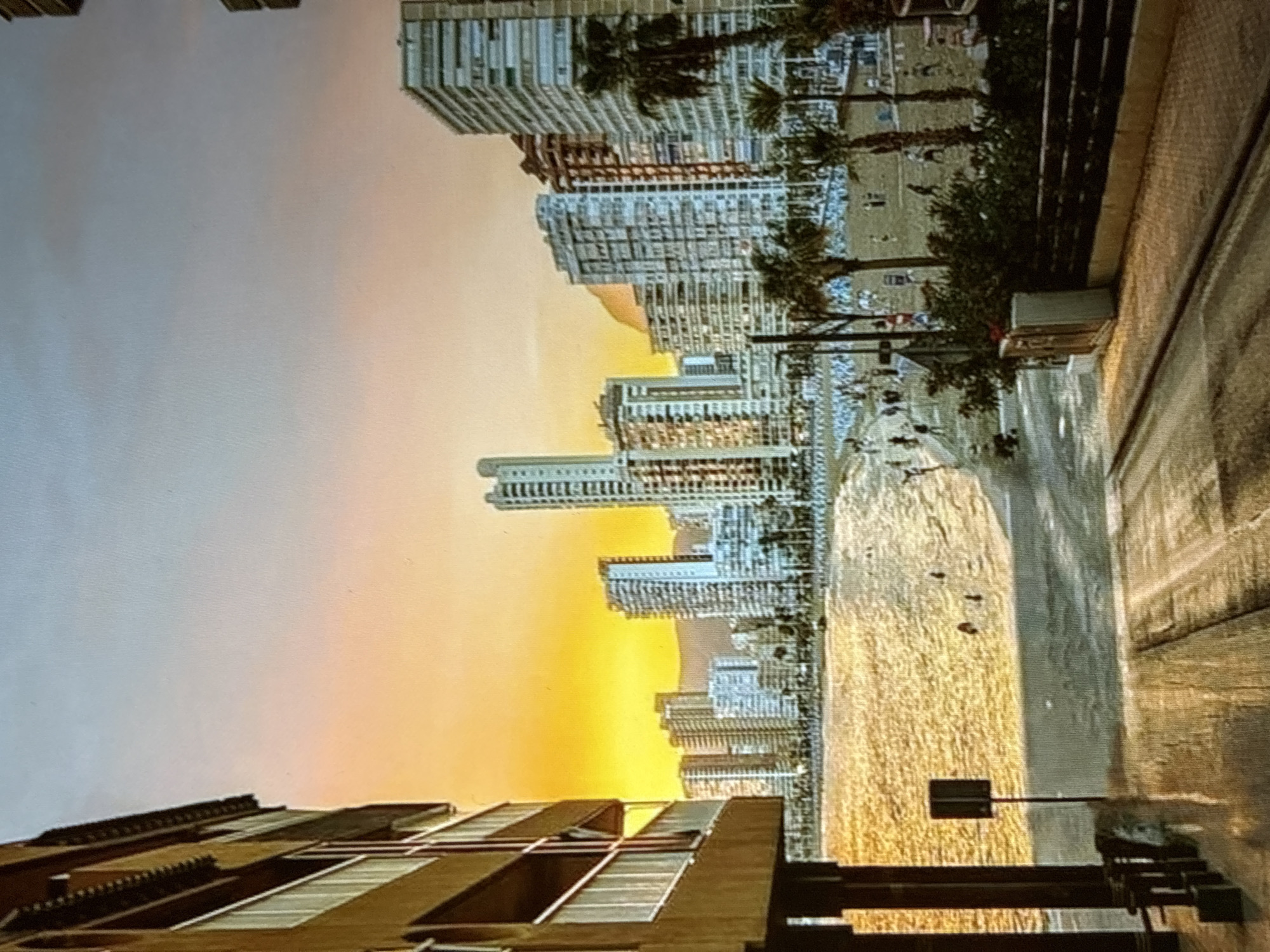 The image depicts a coastal city skyline at sunset. The sky transitions from a bright yellow near the horizon to a pale blue at the top. A cluster of tall buildings, characteristic of a modern city, lines the waterfront, with some appearing to be hotels or apartment complexes. Palm trees are visible in the foreground, suggesting a warm climate. The beach below is populated with numerous small figures, likely people enjoying the evening. In the immediate foreground, there is a structure that resembles a balcony or terrace of a building, and a paved pathway or road. The overall impression is a vibrant urban beach scene during the golden hour.
The location is not Pori, Finland, as Pori is an inland city with a coastline on the Gulf of Bothnia, but it is not known for the type of densely packed, high-rise development seen here nor for palm trees. This type of architecture and vegetation is more characteristic of Mediterranean or other warm coastal regions.