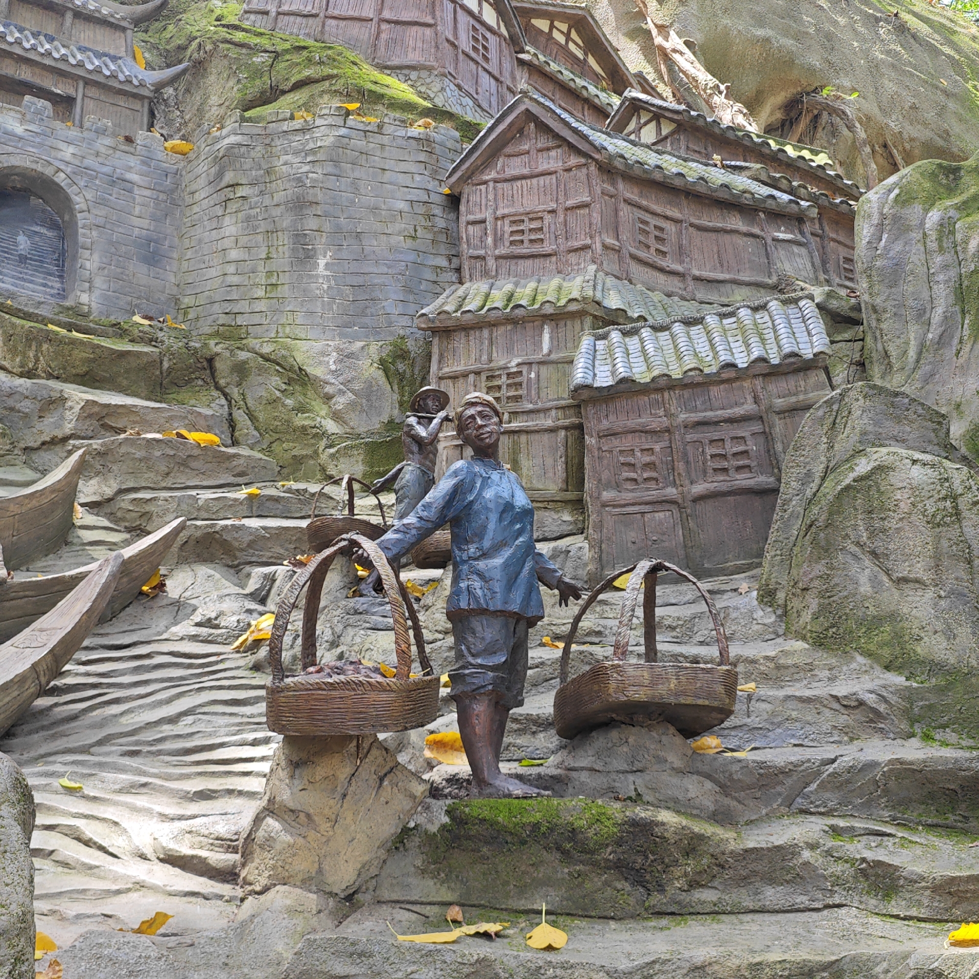 The image depicts a diorama of a Chinese village scene, likely a tourist attraction or public art installation. It is set on a rocky, moss-covered hillside. The architectural style suggests traditional Chinese design.
In the foreground is a bronze statue of a man carrying two woven baskets suspended from a yoke. The baskets appear to contain goods. Another statue of a man carrying the same yoke is behind him and slightly up a set of stairs.
To the left of the statues, a miniature stone wall with an arched opening is seen. To the right and behind the statues are miniature wooden buildings.
The setting appears to be outdoors with natural rock formations integrated into the diorama. The color palette is muted, with grays, browns, and greens dominating. Scattered yellow leaves suggest an autumn season.
It is likely the location is in the Yuzhong District, China, which is known for its hilly terrain.
