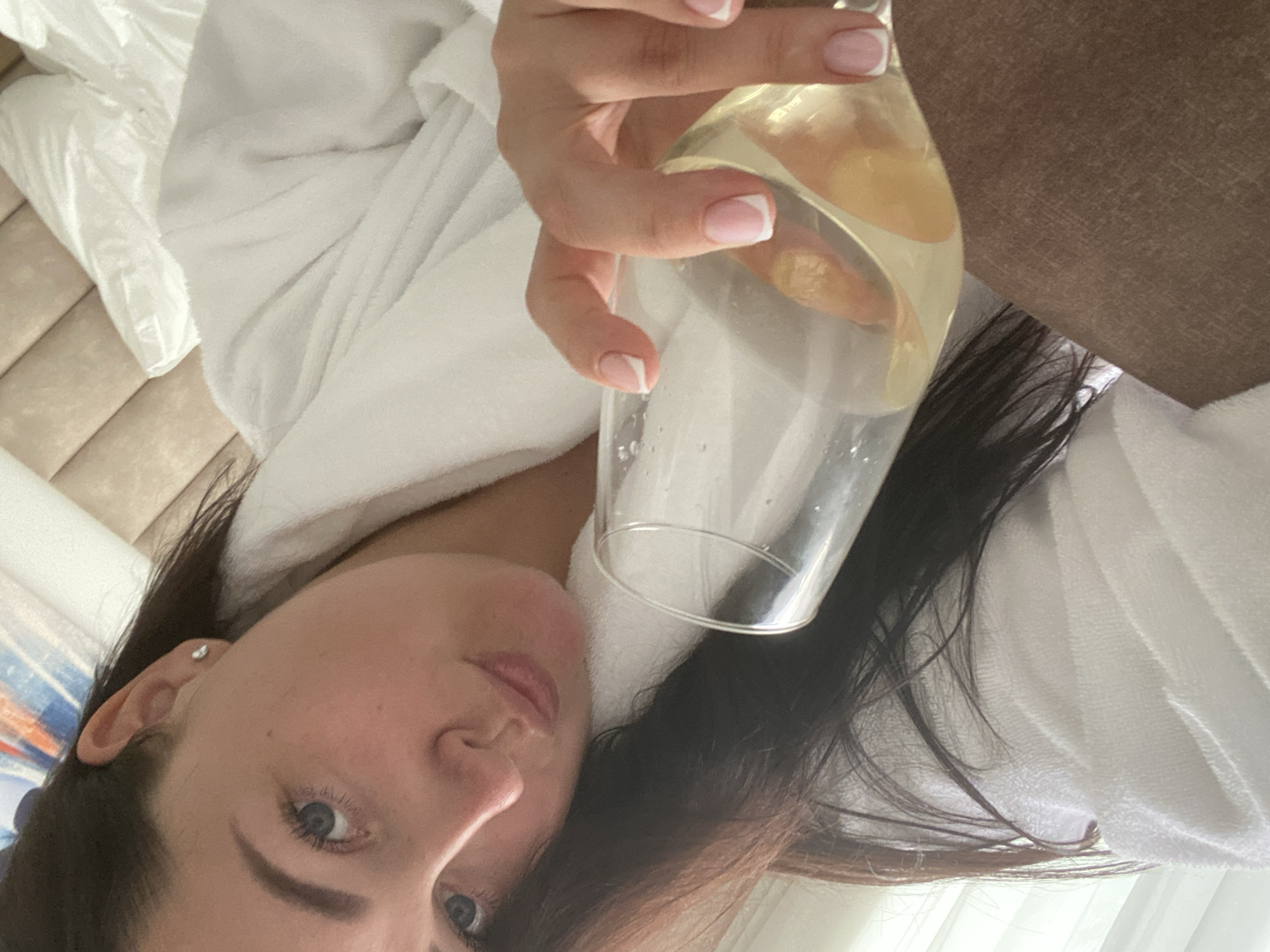 A woman with long, dark hair and blue eyes is lying on her back, wearing a white bathrobe. She is holding a glass of a light-colored liquid, possibly wine, with a piece of fruit in it. Her nails are manicured with a white French tip. The background includes a bed with light-colored linens and a headboard, as well as a curtain. The scene may be set in a hotel room in Sochi, Russia, based on potential tourist imagery.