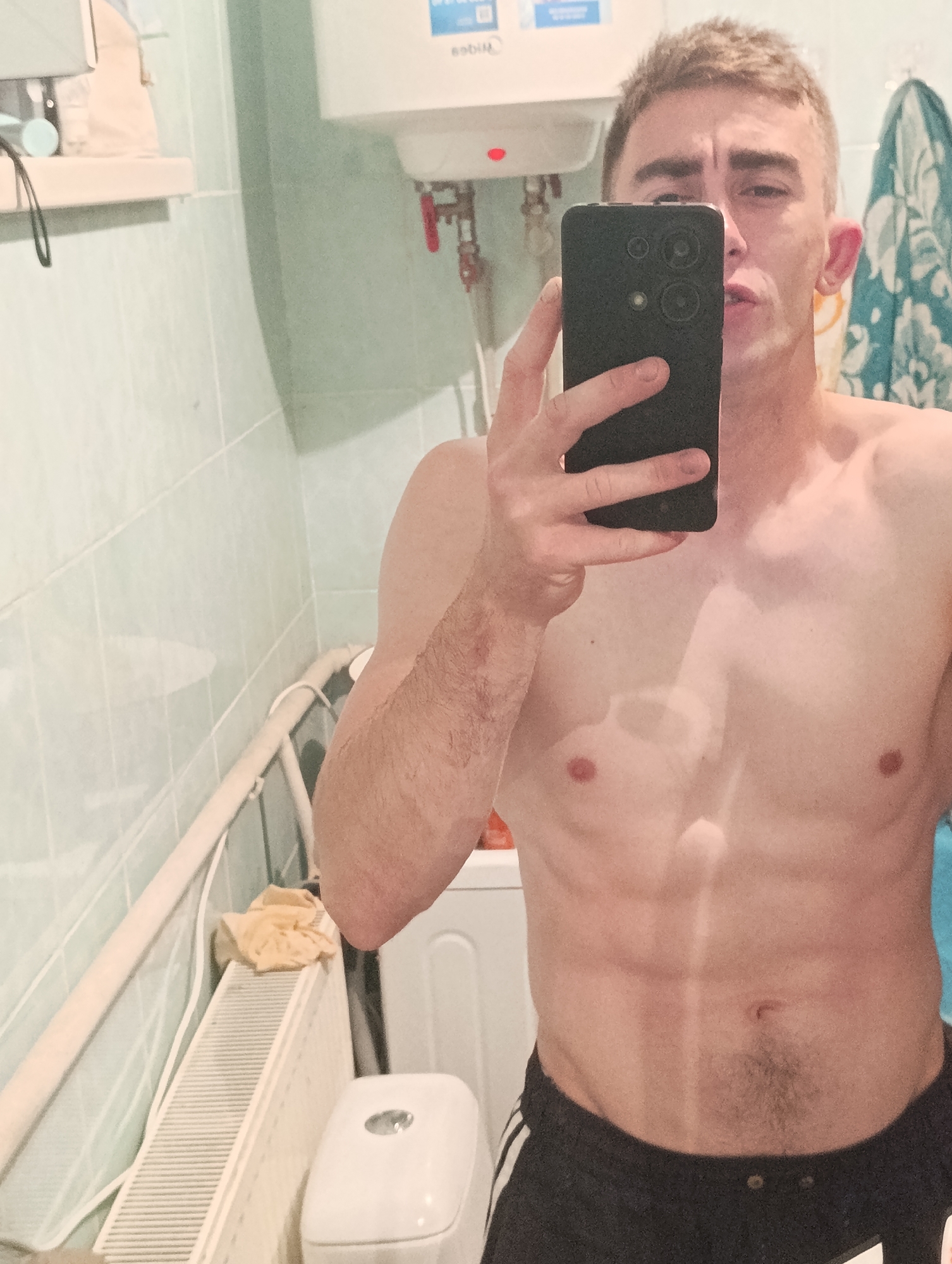 A shirtless young man with short, light brown hair takes a mirror selfie in a bathroom. He is wearing black athletic shorts with white stripes. His abdomen is visible and appears toned, with a distinct line down the center. The bathroom has light blue tiled walls and a white radiator with a yellow cloth draped over it. A white toilet is partially visible. A white water heater is mounted on the wall above the tiled area. A black smartphone is held in his right hand, obscuring part of his face and chest. The image appears to be taken in Fastiv, Ukraine.