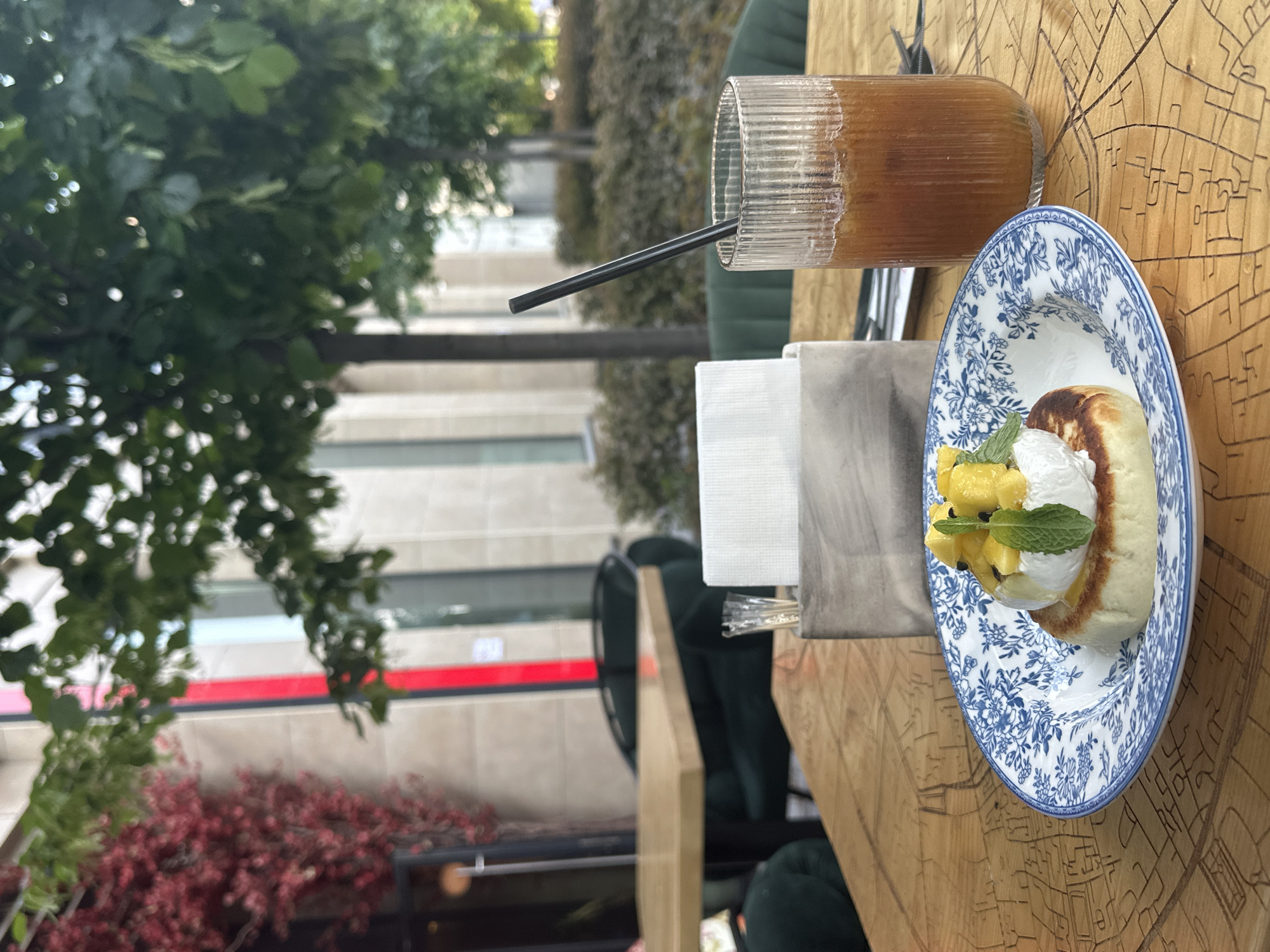 The image shows a close-up of a table at an outdoor cafe in Novosibirsk, Russia. A wooden table has a meal and a drink placed on it. A dessert, possibly a pancake or syrniki, topped with whipped cream, diced pineapple, and mint leaves, is served on a blue and white patterned plate. Next to it is a tall glass filled with an iced beverage, likely coffee or tea, with a black straw. In the background, there are lush green trees and foliage, suggesting a pleasant daytime setting, possibly spring or summer. The architecture in the background, with its large windows and red and white detailing, hints at a modern building. The overall ambiance is relaxed and inviting, typical of a cafe environment. The wooden table has an etched design that resembles a city map. There is no visible text in the image.