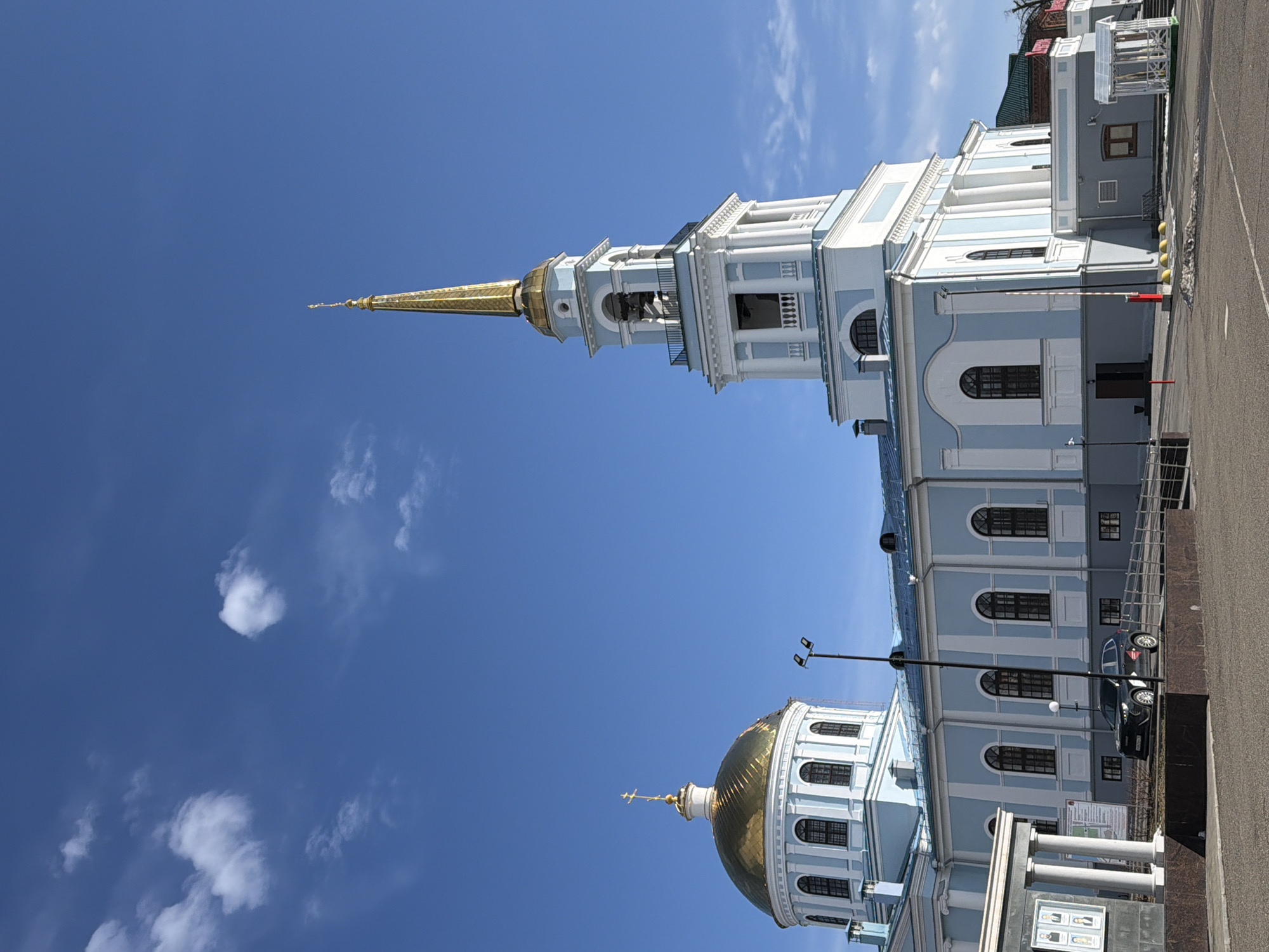 The image depicts the architectural structure of the Annunciation Cathedral located in Votkinsk, Russia. The cathedral is characterized by its light blue and white color scheme, featuring two prominent golden domes and a tall steeple. The clear blue sky is visible in the background, with a few scattered clouds. Adjacent to the cathedral is a parking area with a car visible, as well as a walkway and possibly other commercial buildings.
