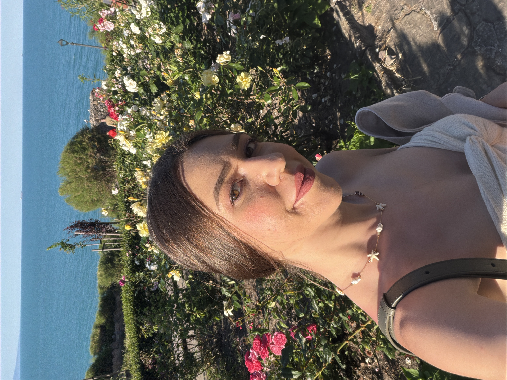 A young woman with long brown hair pulled back is captured in a close-up selfie, her face turned towards the camera. She is wearing a light-colored, possibly off-the-shoulder top and a delicate necklace with shell and starfish charms. Her makeup includes blush, eyeshadow, and lipstick. The background reveals a vibrant garden with numerous pink and yellow roses in bloom. Beyond the garden, a stone wall separates it from a lush green hedge, which then gives way to the clear blue sea and a distant, hazy shoreline with faint structures. The setting sun casts a warm glow across the scene, highlighting the textures of the flowers and the woman's skin. The image appears to have been taken in Sozopol, Bulgaria, likely in a scenic location overlooking the Black Sea, possibly within the Old Town or a coastal garden.