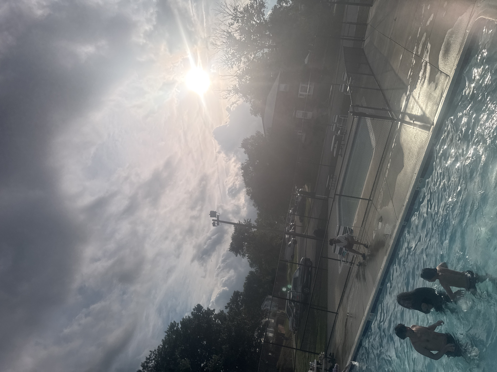 The image shows an outdoor swimming pool. Three people are in the water. Another person is sitting on a chair near the edge of the pool. A chain link fence runs along the side of the pool. Beyond the fence, there is a parking area with parked cars, trees, and a building. The sky is cloudy with the sun visible. The location is Unknown, Unknown.