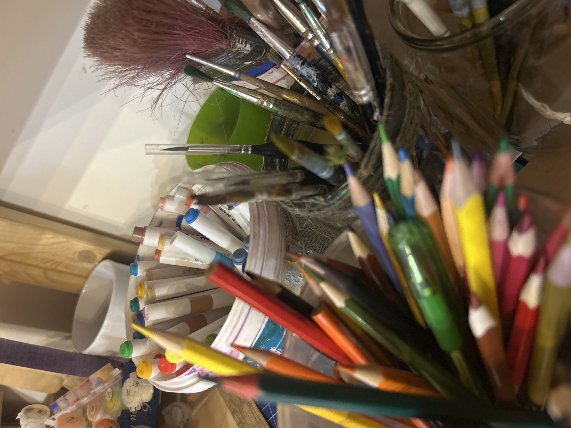 The image is a close-up, top-down shot of an artist's workspace. The scene contains a collection of art supplies including paintbrushes in a glass jar, colored pencils and tubes of paint arranged in a circular manner. The paint tubes vary in color and are arranged on a palette. The context and details provided do not indicate any identifiable location in Izhevsk, Russia, or any people, actions, or interactions. The photograph presents a still life focused solely on the artist's materials.