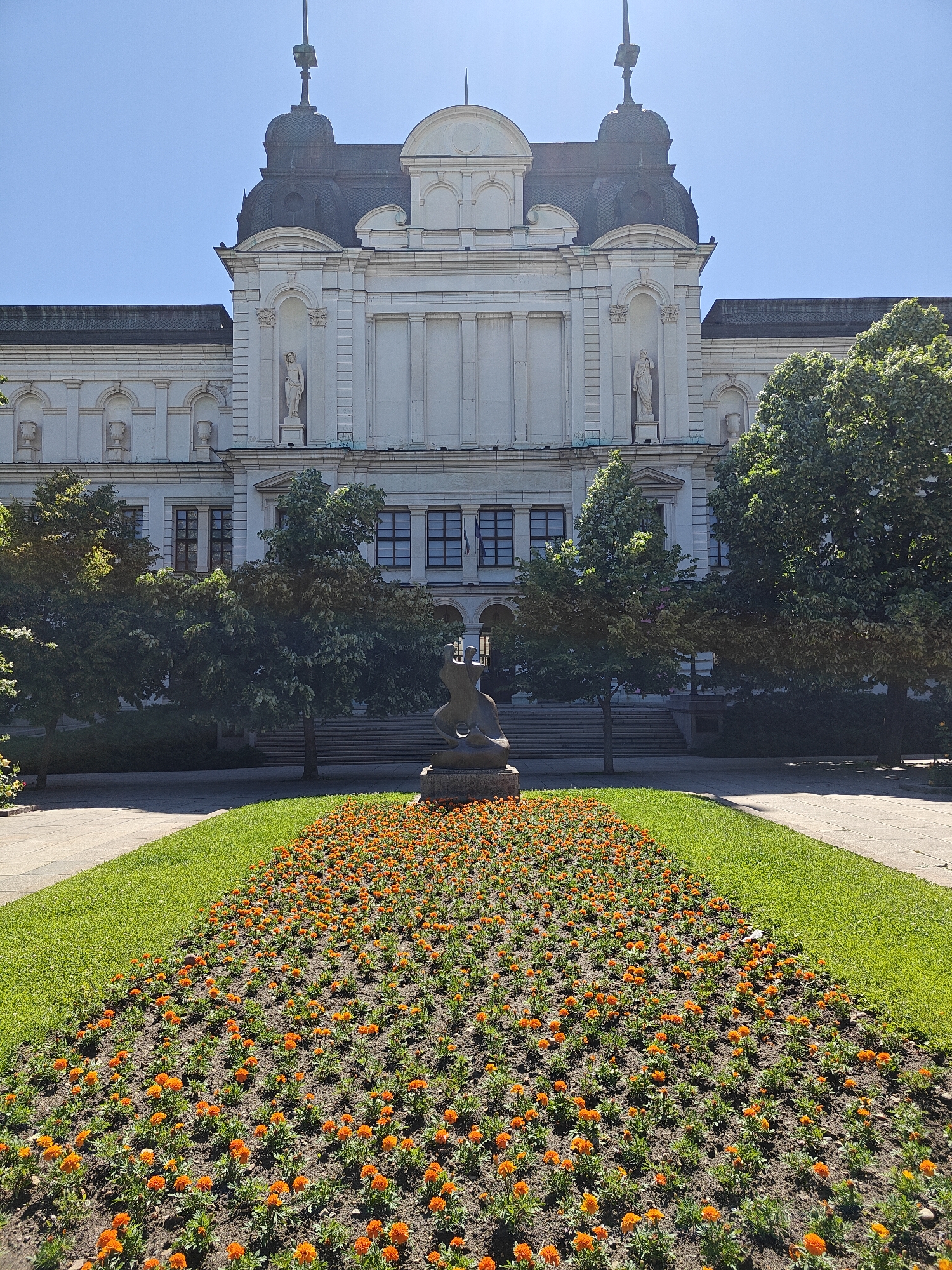 A full outdoor shot depicts the National Art Gallery in Sofia, Bulgaria, on a bright day with clear blue skies. The gallery is a stately, white-colored building with a dark roof, featuring classical architectural elements like statues in niches and symmetrical design. A few trees are visible in front of the building, partially obscuring the main entrance. In the foreground, a vibrant flower bed of orange blooms stretches out from the statue. A paved walkway flanks the flower bed on both sides, leading towards the gallery entrance. The overall scene is well-maintained and composed, capturing the beauty of the gallery and its surroundings.