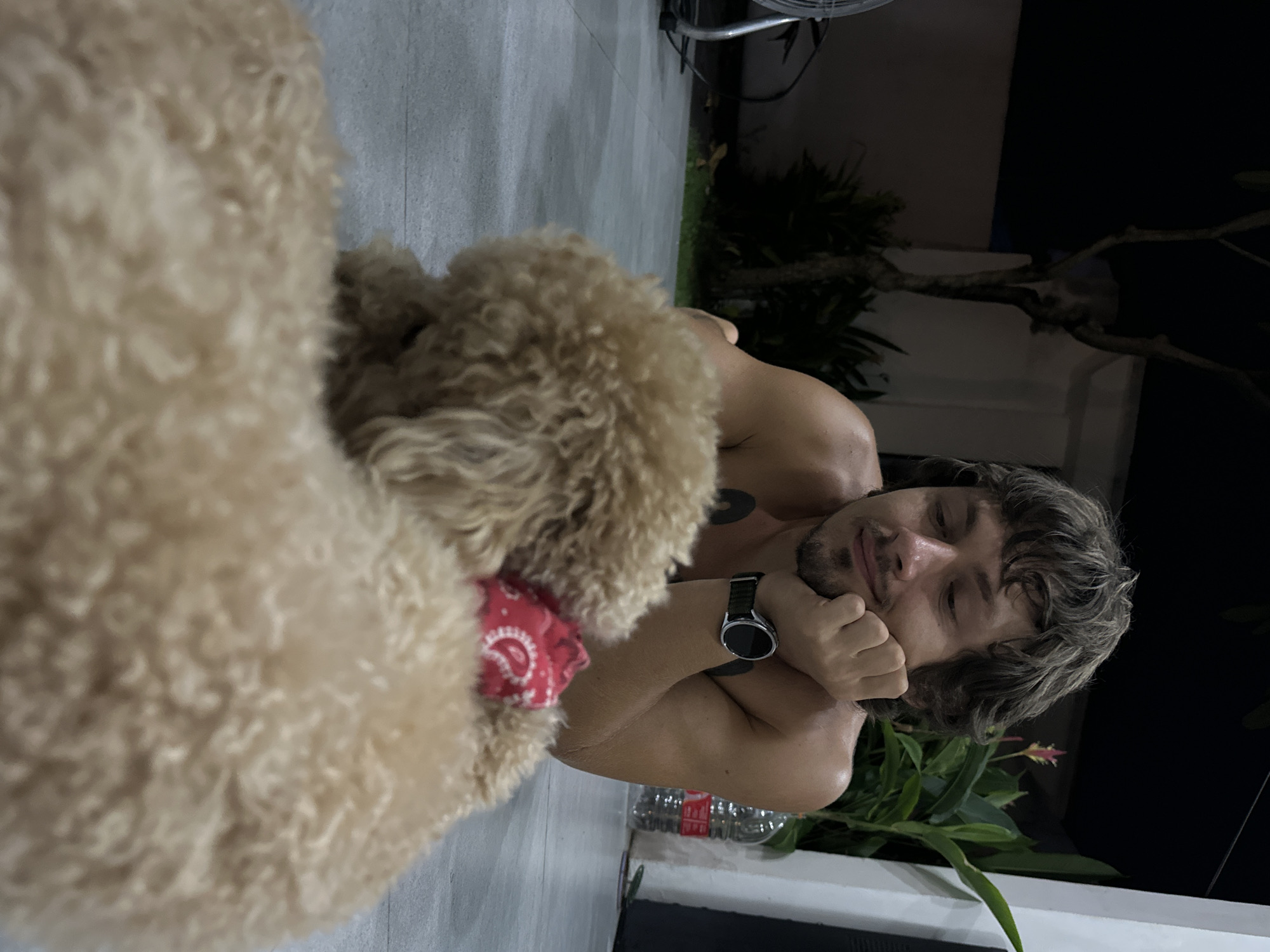 A man with fair skin and light brown hair is lying on a gray tiled floor next to a golden doodle dog wearing a red bandana. The man is resting his head on his hand, wearing a black smartwatch on his left wrist. A half-empty water bottle and green plant are near him. The environment suggests an outdoor patio or deck, with greenery and dark foliage in the background, characteristic of the location in Dalung, Indonesia.