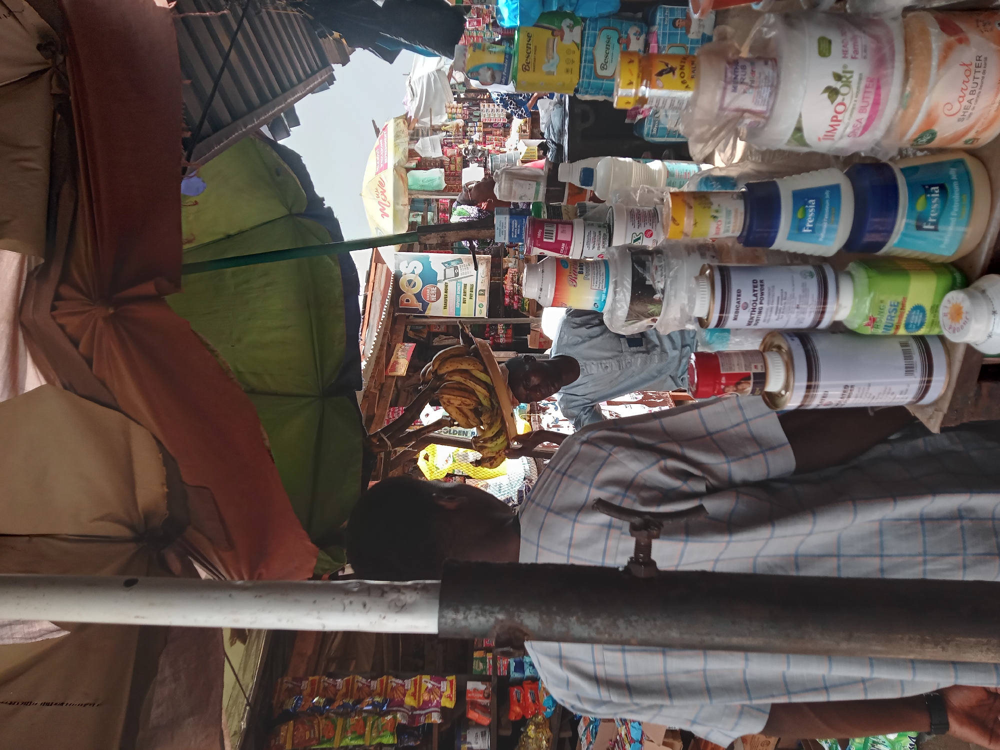 The image shows a bustling open-air market in Sabon Tasha, Nigeria. A man wearing a checked shirt stands in the foreground, partially obscuring the view. Behind him, various goods are displayed for sale on makeshift shelves, including personal care products like "Fressia" and "IMPO-ORI" shea butter, "Besense" baby powder, and medicated mentholated dusting powder. A sign advertising POS (Point of Sale) services is visible. Vendors and customers are discernible in the background, shaded by colorful umbrellas. There are also bananas hanging above a small wooden table.