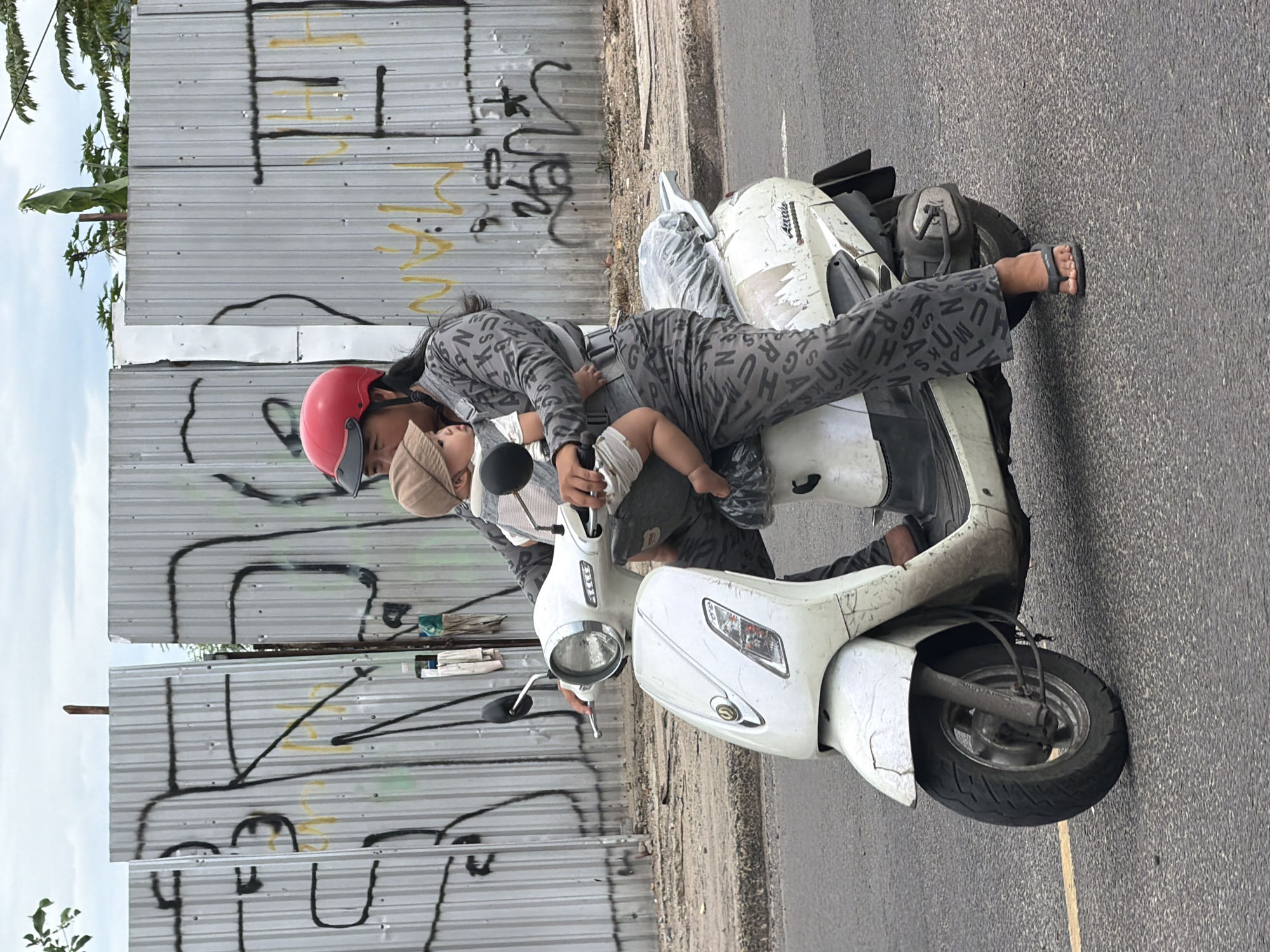 A woman wearing a red helmet and a gray outfit with a pattern of letters rides a white scooter on an asphalt road. She is carrying a baby in a gray carrier. The baby is wearing a light brown hat. Behind them is a corrugated metal fence covered in graffiti. The scene appears to be in Nha Trang, Vietnam, based on the presence of similar fences and typical street scenes. The road has a yellow line marking. The sky is overcast.