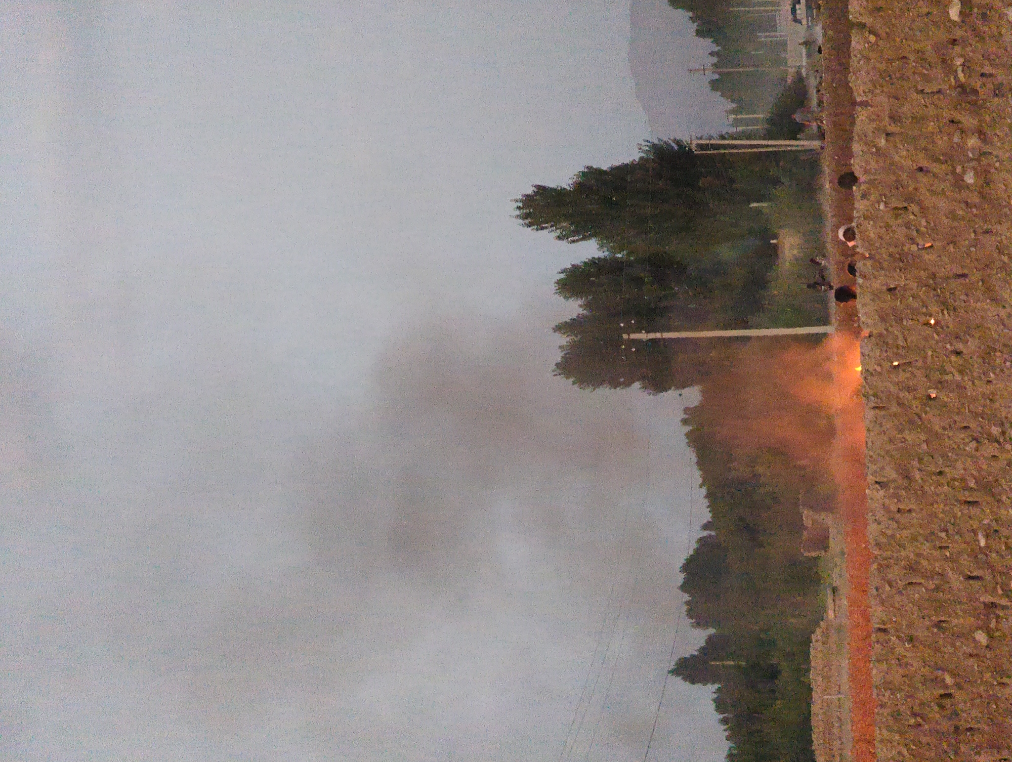 In an outdoor scene in the village of Uch-Dobo, Kyrgyzstan, a fire burns in the foreground, creating orange flames and smoke. The fire is situated on a dirt or gravel surface behind a textured, brown barrier that appears to be a wall or embankment. Beyond this, a thin orange band runs horizontally across the middle ground, potentially indicating more burning material or a different type of ground cover. Trees, some tall and dark green, others appearing more sparse, are visible behind the fire, suggesting a rural or semi-rural environment. A few indistinct human figures are present, some near the fire and others further back. Several utility poles and power lines are present in the mid-ground, indicating the presence of infrastructure. The sky is overcast and a muted grey. In the far background, hills or mountains are dimly visible.