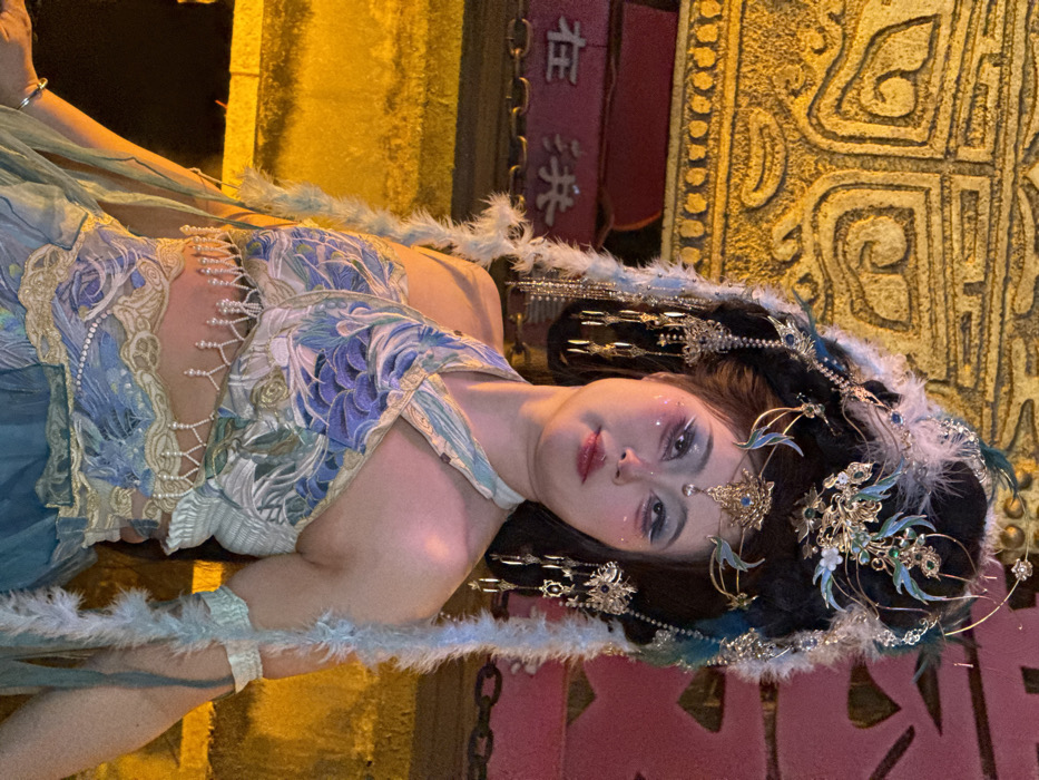 A young East Asian female is centrally depicted, posed in elaborate traditional-style attire against a warmly lit architectural background. Her dark hair is intricately styled into a voluminous updo adorned with a prominent gold and teal headpiece, featuring feathers, metallic scrollwork, blue gemstones, and dangling chains with pearl-like accents. Long, light blue feathery decorative strands cascade from her hairstyle. Her face features dramatic makeup, including pale foundation, extensive blue eyeshadow with glitter, prominent dark eyeliner, pink blush, and rosy lips, with small decorative elements near the outer corners of her eyes.
She wears a two-piece ensemble: a halter-style crop top with a cross-front design, patterned in shades of blue, green, and gold with scale or feather motifs. A fringe of white pearl beads hangs from the bottom edge of the top, exposing her midriff. Below the crop top, a decorative band with gold and blue patterns is visible, leading to a flowing lower garment or skirt in varying shades of blue and green fabric. A light blue feathery armband is present on her left upper arm, and a simple silver bracelet is on her left wrist. She gazes slightly upwards and to her left, displaying a subtle smile.
The background consists of ornate, traditional Chinese architecture. To her left and above, a large, golden-yellow illuminated panel with raised, textured patterns is visible. Below this panel, partially visible Chinese characters "在 洪" (Zài Hóng) are present on a red surface. The scene is located within the Hongyadong complex in Yuzhong District, Chongqing, China, identifiable by the distinctive architectural style and the visible characters which are part of the complex's name.