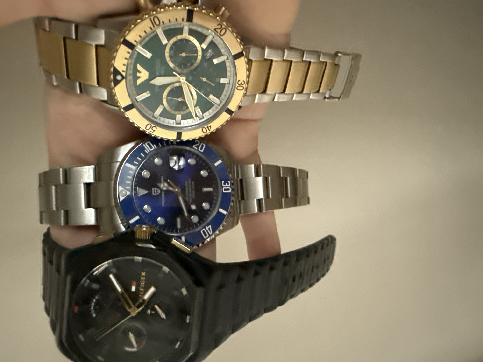A close-up, overhead shot displays three wristwatches worn by an individual. From left to right, the watches are: a black Tommy Hilfiger watch with a textured strap and gold accents; a silver and blue dive watch with a rotating bezel and a dark blue dial; and a gold and silver watch with a green dial and a gold bezel, featuring the Emporio Armani logo. The watches are held in the foreground against a neutral, light-colored background, likely an indoor setting. The image does not provide any contextual information or visual cues to suggest a location in the city of Burgas, Bulgaria.