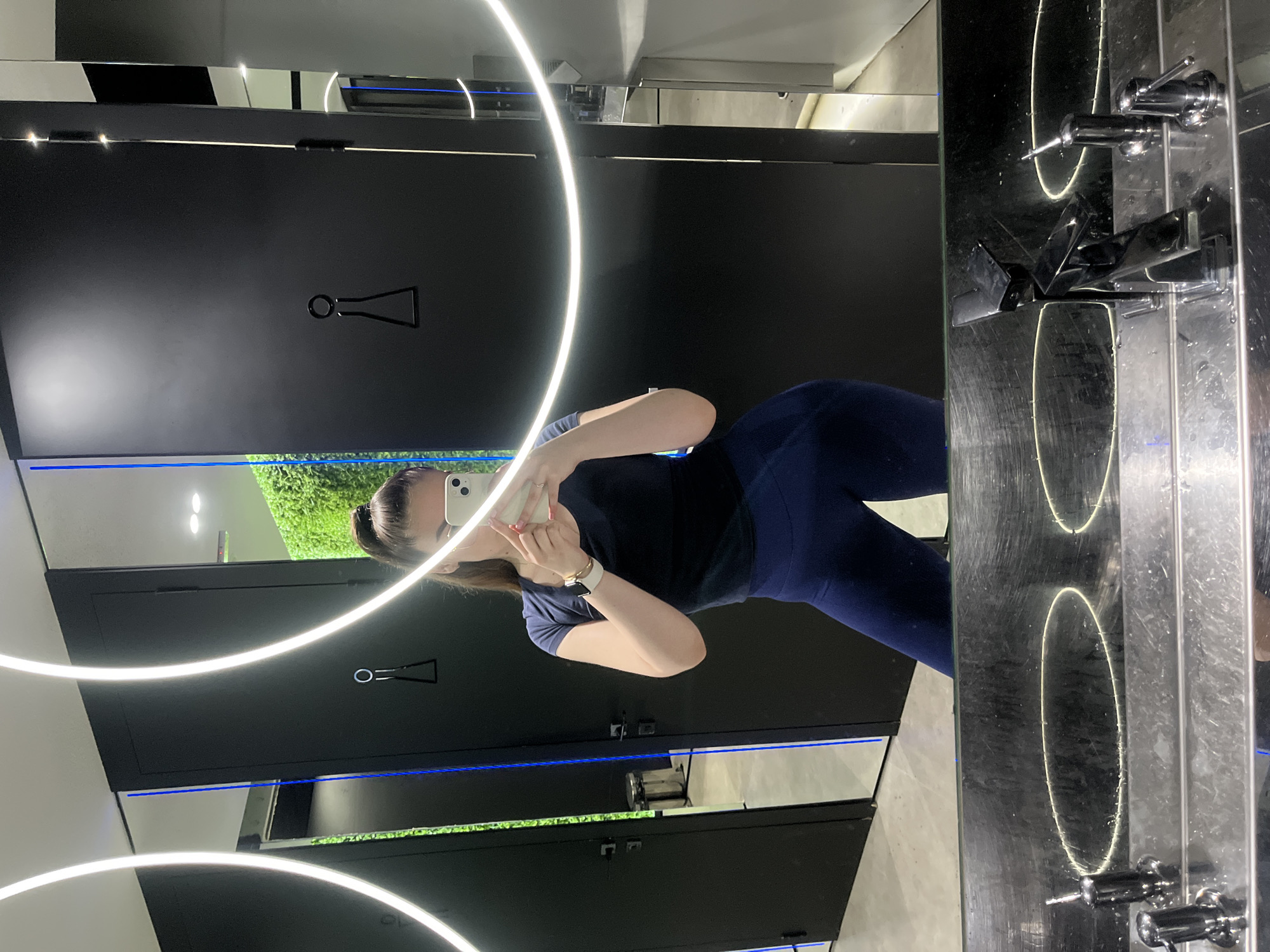 A person stands in front of a mirror in a public restroom in an unknown location. The restroom features black restroom stalls, each with a symbol on the door indicating its purpose. A stripe of green, possibly artificial foliage, is visible behind the stalls. A large, circular light fixture is reflected in the mirror, along with the person taking a selfie. The mirrored wall features chrome fixtures, including a sink and water faucet. The floor is light gray and appears to be tile.