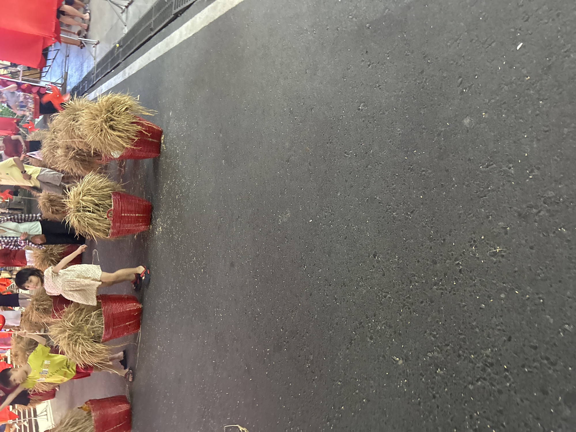 The image shows a street scene in Bắc Nha Trang, Vietnam. There are several red baskets filled with dried, wheat-like grass or stalks lining the side of the road. Children and adults are interacting with the baskets, some standing near them. The street itself appears to be asphalt and is marked with a solid white line along the edge. A red banner or covering is visible in the background on the left side.