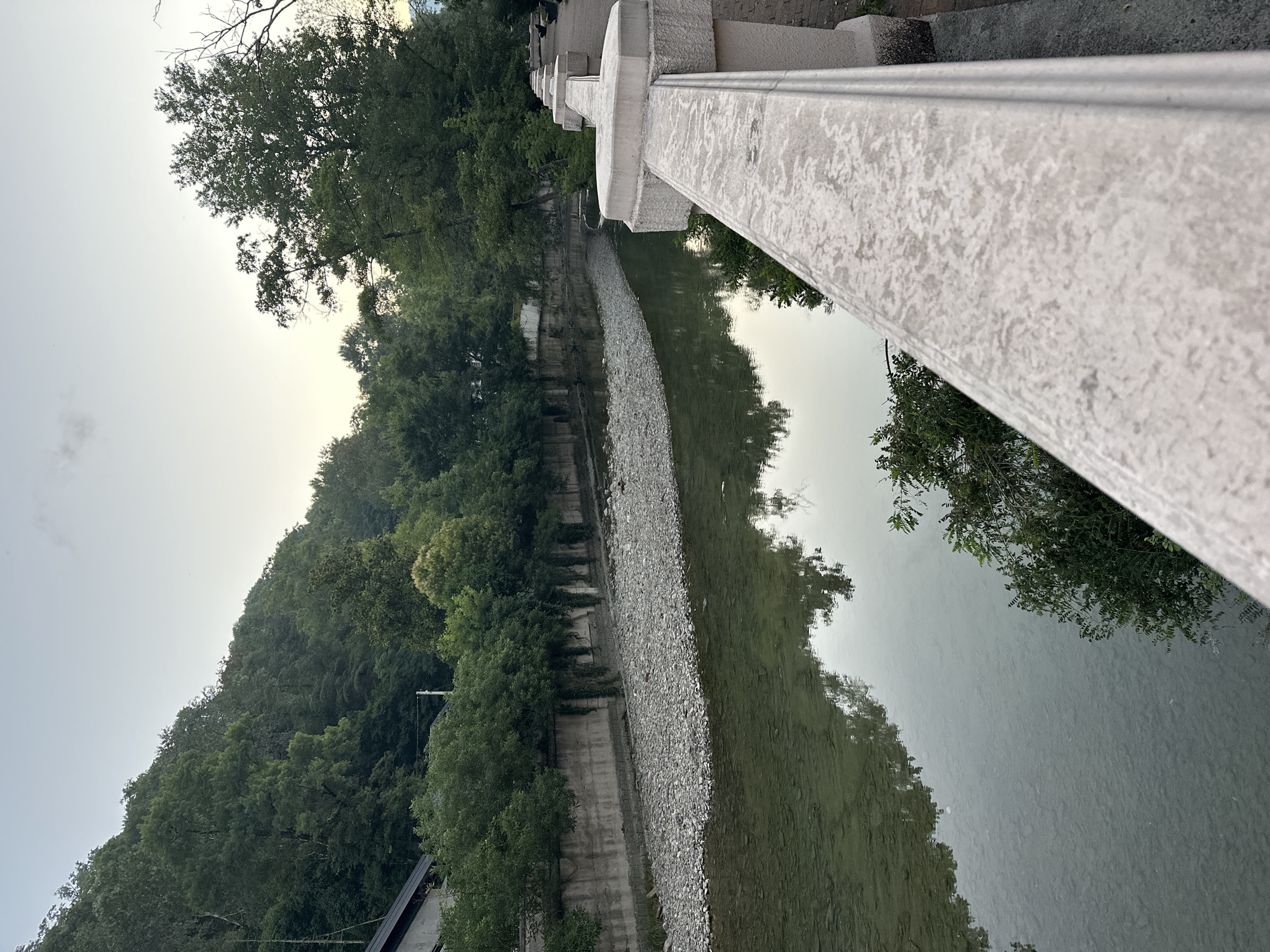 The image depicts a view from a concrete bridge railing, looking out over a river in Sochi, Russia. The river has a pebbly bed and its surface reflects the overcast sky and surrounding lush green trees that line its banks. On the left side of the river, a concrete retaining wall is visible, with some greenery cascading over it. On the right side of the river, a paved walkway can be seen, also bordered by the concrete railing. The scene appears to be a public park or promenade along the river, likely a tranquil spot within the city. The sky is a pale grey, suggesting it might be early morning or late evening. No people are visible in the frame.