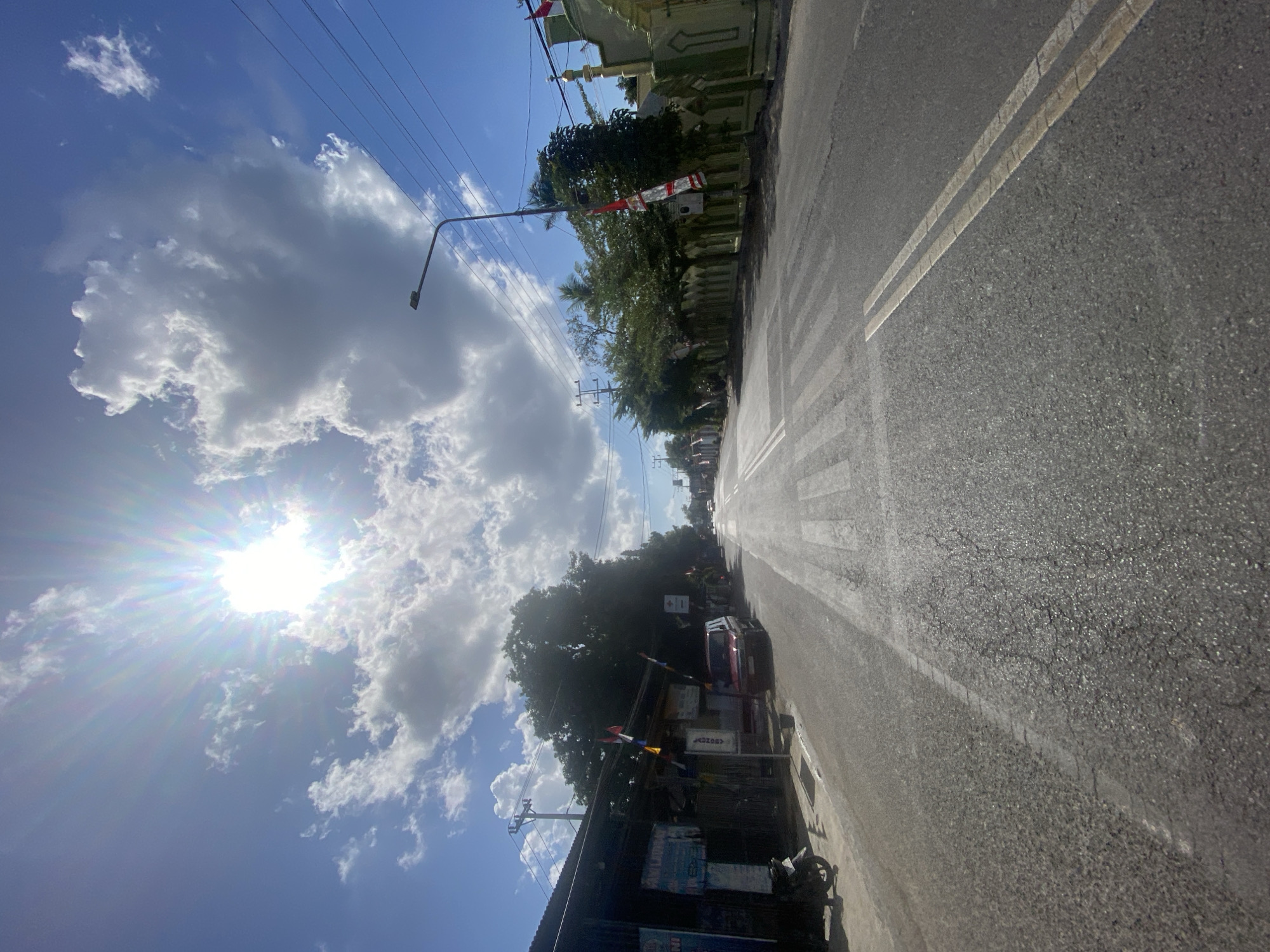 The image depicts a street scene in Bentengnge, Indonesia, under a bright, sunny sky with scattered clouds. The sun is prominently visible, creating a lens flare effect. On the left side of the road, there are low-rise buildings with signage, and a parked motorcycle. The road itself is paved and shows signs of wear. A pedestrian crossing is marked on the road. On the right side of the road, there is a green and white building, possibly a mosque or public building, with decorative elements and flags. Trees line the right side of the street, and there are utility poles with overhead wires. The perspective suggests the photo was taken from a slightly elevated position, looking down the street.