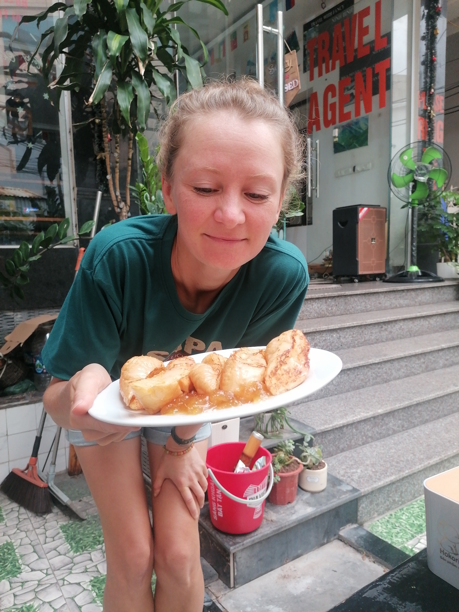 A woman holds a plate of food outside a travel agency on a street in Bai Chay Ward, Vietnam. The woman is fair-skinned with blonde hair pulled back. She wears a dark green t-shirt and light-colored shorts. Her gaze is downward toward the plate, which contains pieces of pastry drizzled with a translucent, yellow sauce. Behind her, a travel agency with "TRAVEL AGENT" printed in bold red letters is visible on the glass front door. A green fan, a speaker, potted plants, and a flight of stairs are situated in front of the building's entrance. To the woman's right, a red bucket filled with miscellaneous items and small potted succulents sit on a ledge. The ground is tiled with gray and green pavers. A broom leans against the wall to the left.