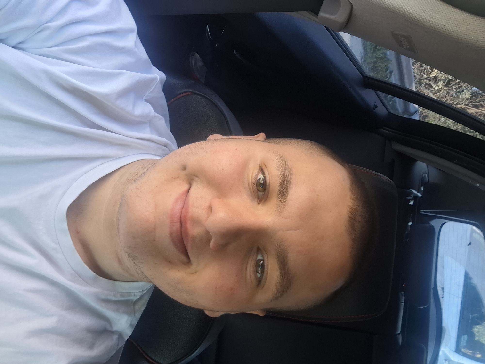 A person is sitting in a car. The person has short brown hair, brown eyes, and is wearing a white t-shirt. The car's interior is black. Outside the car window, there are trees and a building. The image was not captured in the city of Varna, Bulgaria.