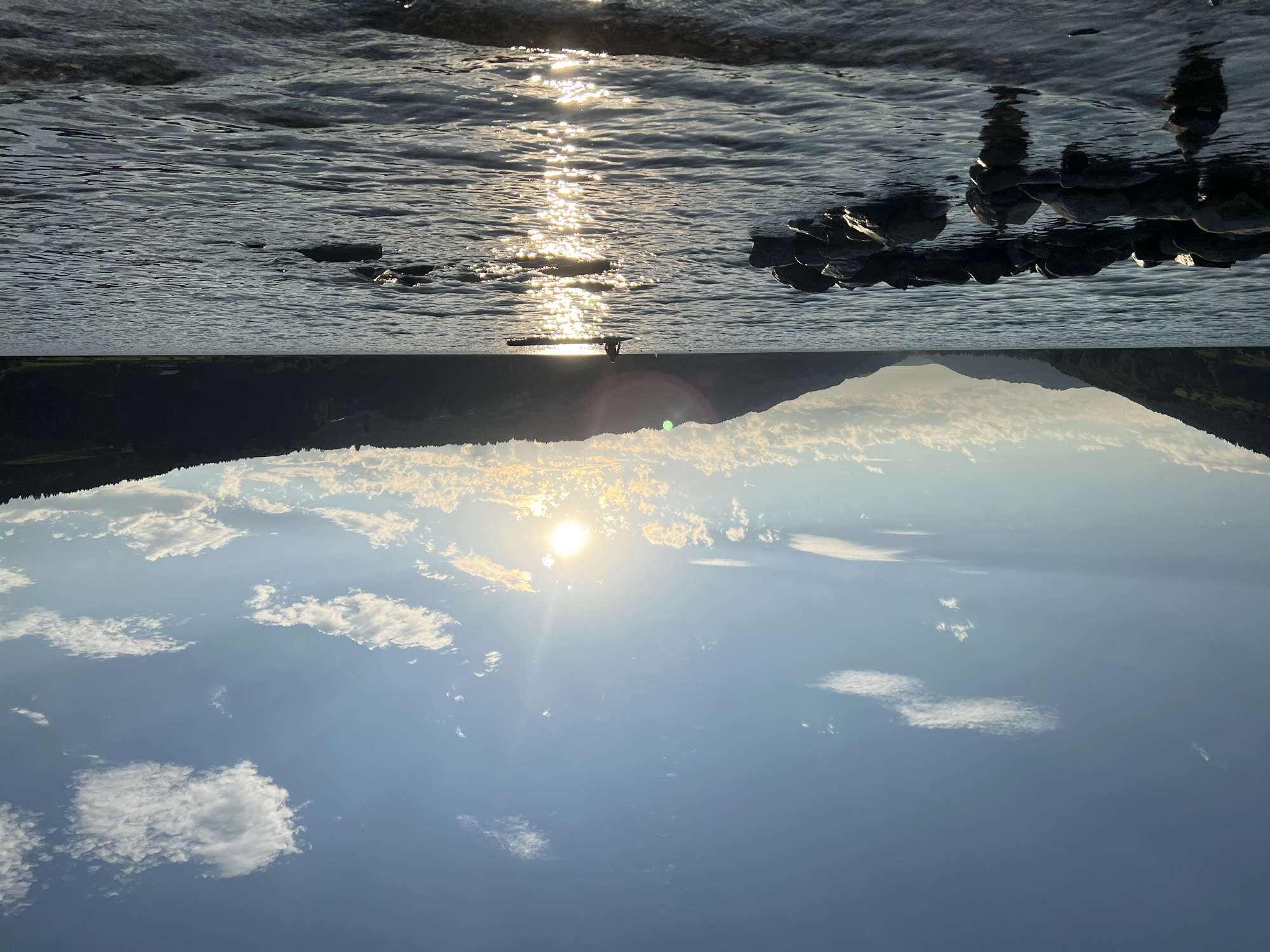 The image depicts a serene lake scene at sunset, likely located near Immenstadt im Allgäu, Germany, given the characteristic Alpine foothills. In the foreground, a ring of dark, wet stones is partially submerged in the rippling water. Further out on the lake, a lone individual stands on what appears to be a paddleboard, silhouetted against the setting sun. The sun is low in the sky, casting a brilliant, shimmering reflection across the water's surface. The background is dominated by dark, tree-covered hills that recede into the distance, their slopes softened by the fading light. The sky above is a gradient of blue, with scattered white clouds catching the warm hues of the sunset.