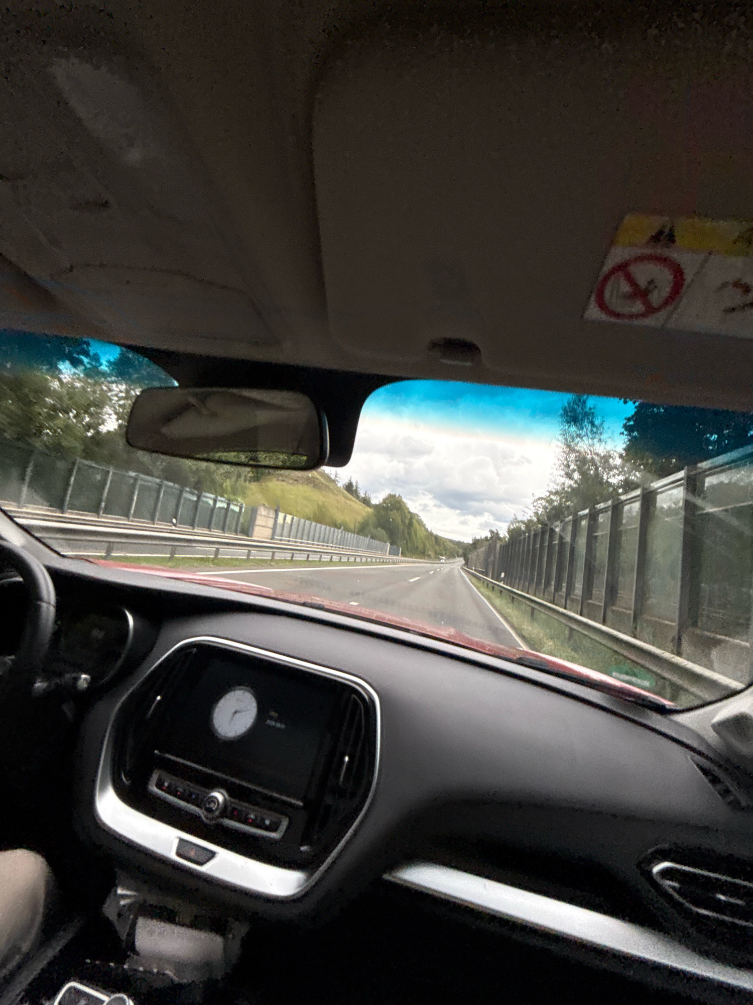 The image, captured from inside a vehicle, shows a view of a highway with sound barriers along the side. The car's dashboard is visible in the foreground, featuring a central infotainment screen displaying a clock and radio controls. The windshield offers a view of the road ahead, flanked by trees and a cloudy sky. There is a sun visor in the upper part of the frame with an airbag warning sticker. The vehicle appears to be in motion, indicated by the blurred scenery and the perspective of the road. The presence of sound barriers suggests this is likely a highway passing through a populated or environmentally sensitive area. No specific landmarks or details pinpoint the location to Herzberg, Germany, beyond the provided context.
