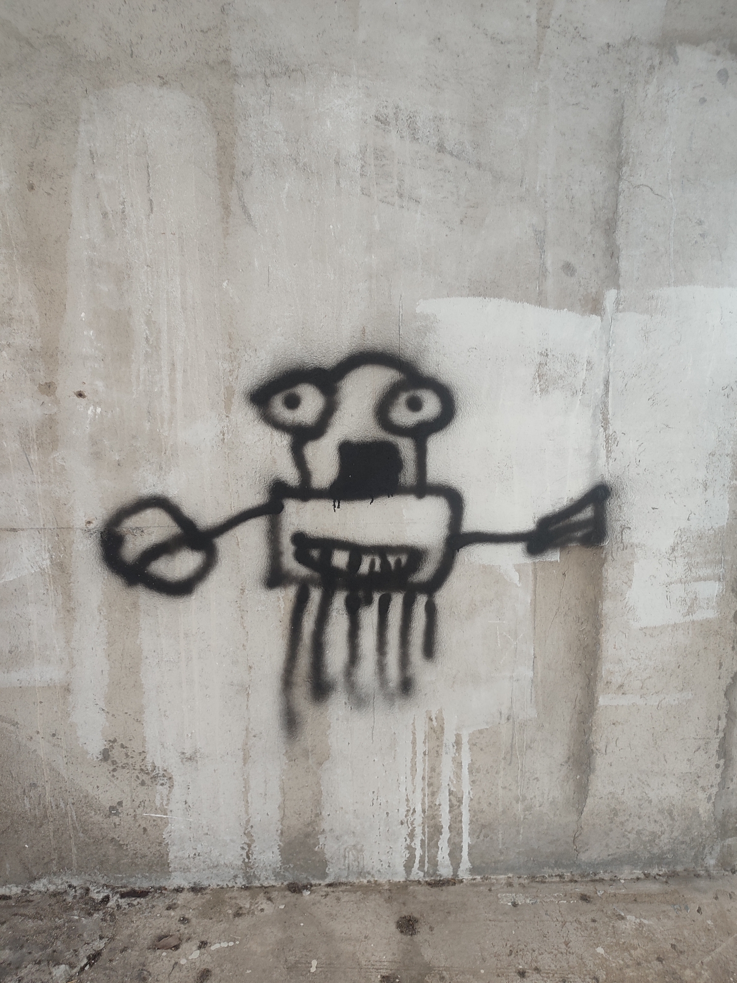 A black spray-painted drawing of a cartoonish figure is on a weathered, light-gray concrete wall. The figure has large, circular eyes, a rectangular body with vertical lines hanging down, and arms extending to the sides, ending in a circle on the left and a triangle with lines on the right. The wall appears to be part of a building or structure, and the bottom edge of the frame shows the beginning of the floor. The image was taken in Nha Trang Ward, Vietnam.