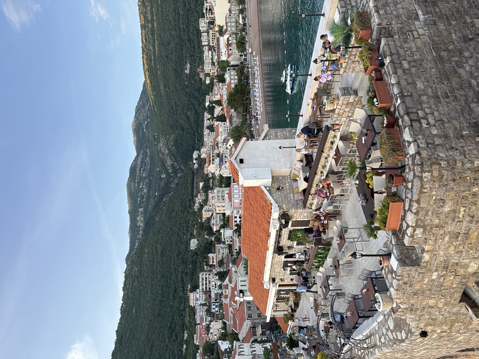 An elevated, angled view captures the coastal town of Petrovac centar, Montenegro. Lush, green mountains rise on the left. The town is a collection of buildings, many with red-tiled roofs, clustered between the mountain and the waterfront. A sandy beach runs along the coast with boats in the water. A stone wall, lined with potted plants and ornate light posts, runs along the right side of the image. Tables and chairs occupy a patio along the wall, with people seated.