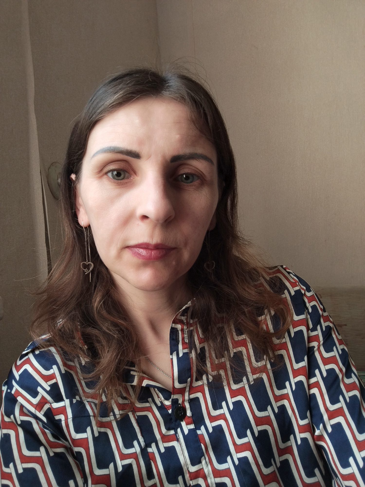 The image shows a medium shot of a woman with brown hair, looking directly at the camera. She is wearing a collared shirt with a geometric pattern of navy blue, white, and red. She has subtle makeup on, including lipstick. She is wearing a long, chain-link earring with a heart charm. The background appears to be a wall with a curtain or drape on the left side. There is no identifiable information in the image that would indicate a specific location in Vinnytsia, Ukraine, or any other city.