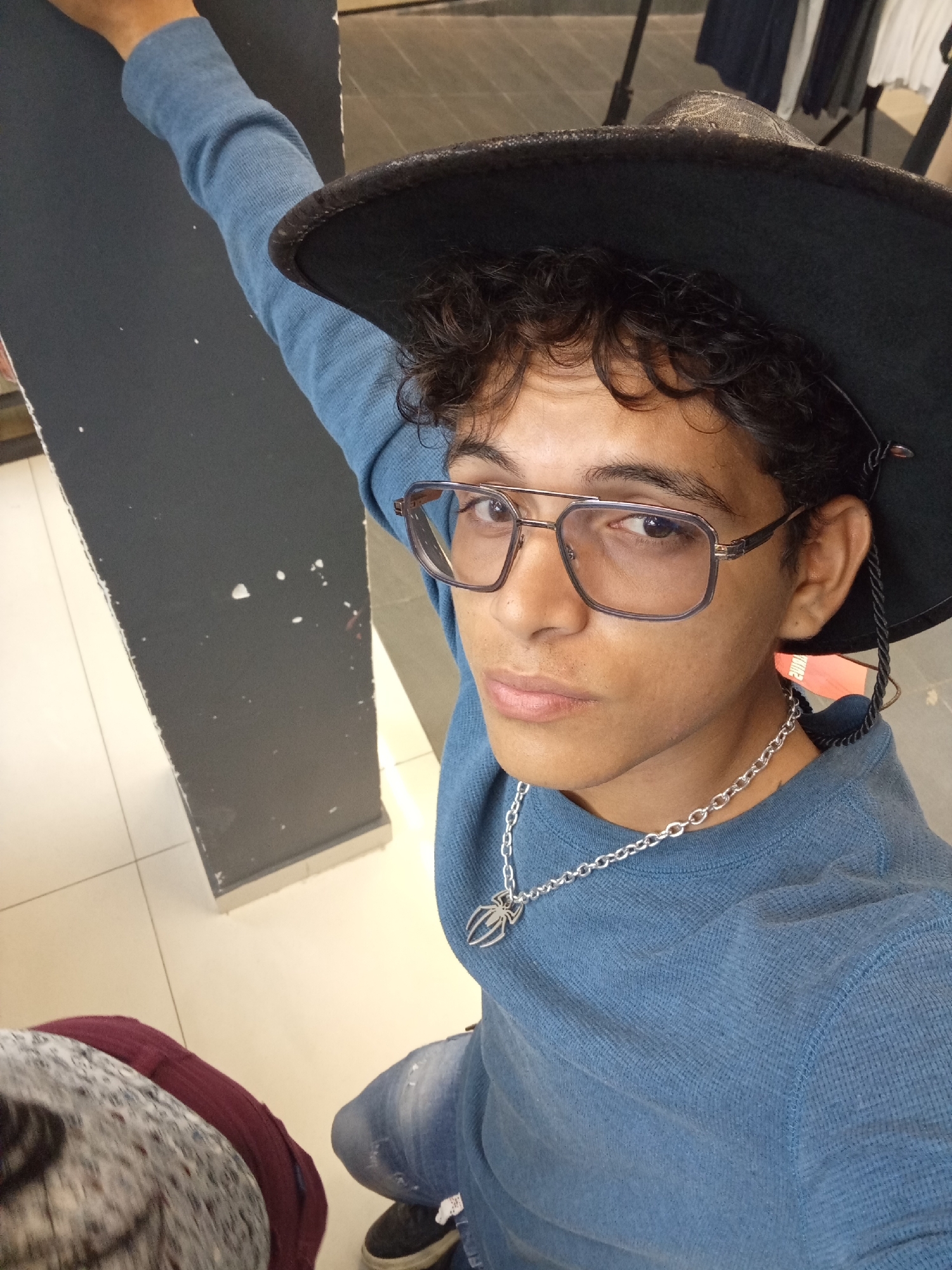 The image presents a young male in an indoor retail setting, consistent with a clothing store. The subject has a medium skin tone, dark curly hair, and wears square-rimmed eyeglasses with a double bridge. He is dressed in a light blue long-sleeved waffle-knit shirt and a dark-colored, wide-brimmed hat, possibly a fedora or cowboy hat. A silver-toned chain necklace with a spider-shaped pendant is visible around his neck. The male looks directly at the camera with a neutral expression. His left arm is extended upwards and left, out of the frame. The background on the left features a dark grey vertical panel or partition with scattered white and faint red marks. The floor is tiled with light-colored squares and dark grout lines. On the right, blurred clothing racks with garments are visible, reinforcing the retail environment. A blurred item, possibly fabric with maroon and grey/white patterns, is in the bottom left foreground. While the environment is clearly a retail store, no visual details within the image directly identify its specific location as the city of Porlamar, Venezuela.