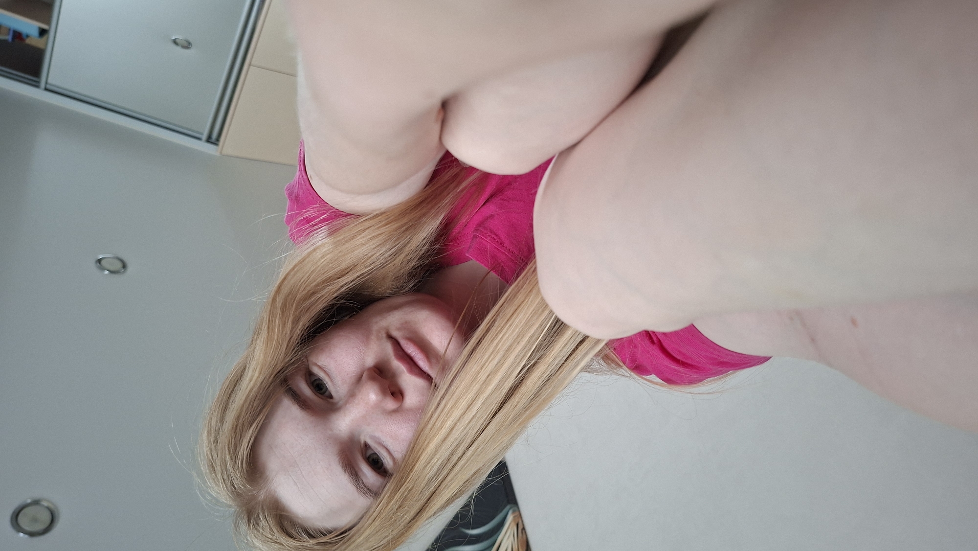 This image depicts a female subject indoors, identified as being in Chaikovsky, Russia. The woman, with long, straight, light blonde hair and fair skin, is wearing a bright pink short-sleeved t-shirt. She is positioned at a low angle, looking directly into the camera with a neutral expression. Her knees and forearms are drawn up towards the camera, occupying the immediate foreground and appearing slightly blurred due to their proximity to the lens. The background features light-colored walls and a matching ceiling, which contains two visible circular recessed light fixtures. To the upper left, a dark, abstract piece of wall art is partially visible. To the mid-right, sections of light-colored cabinetry or a wardrobe with reflective sliding doors can be seen, indicating a domestic interior setting.