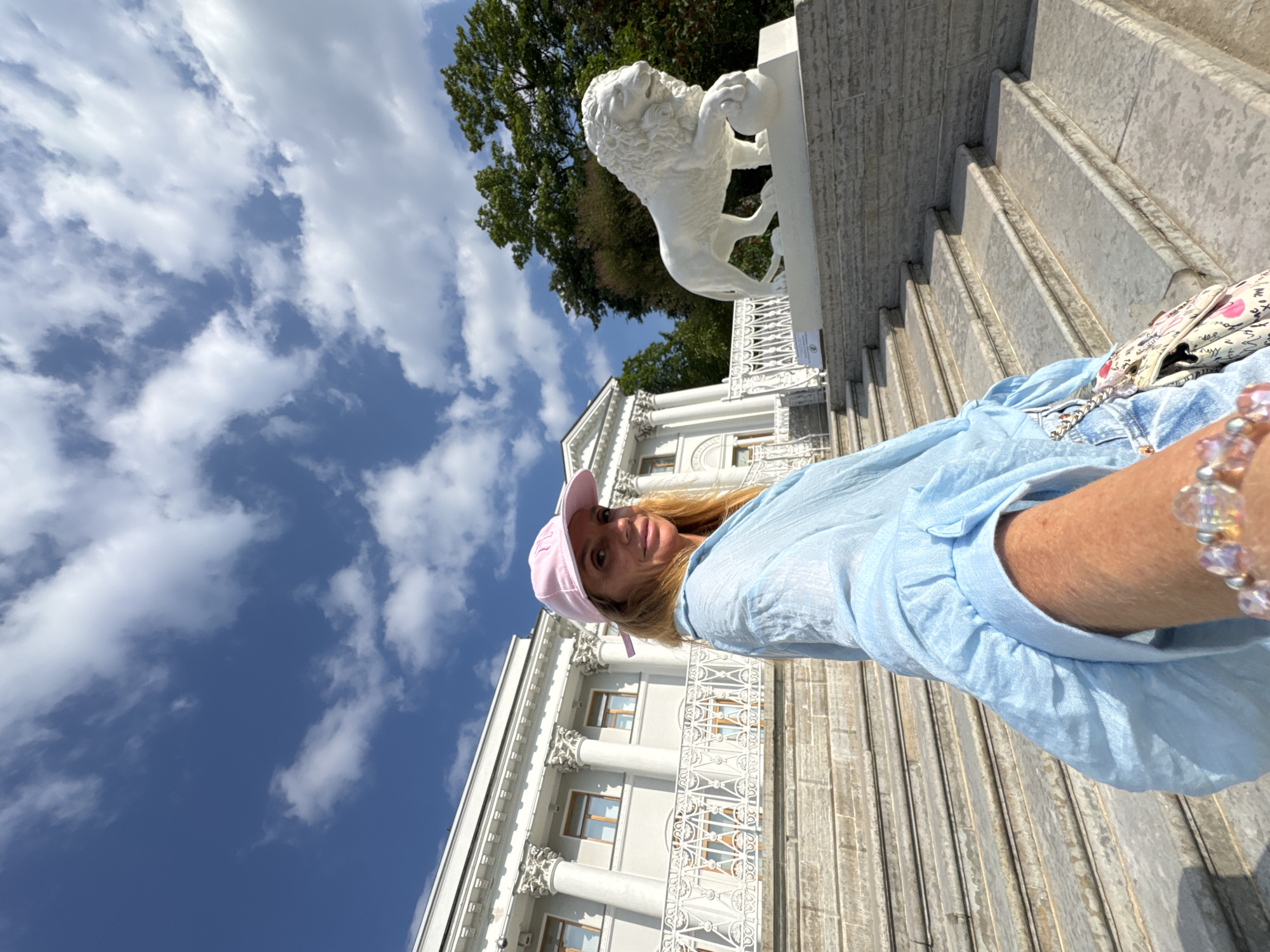 A woman is taking a selfie outdoors, with a large white building with columns and ornate railings in the background. She is wearing a light blue shirt and a pink baseball cap. In the background, on top of the stairs of the building, there is a white sculpture of a lion holding a ball. The sky is blue with white clouds, suggesting it is daytime and the weather is pleasant. The scene appears to be a park or a historical site. The woman is smiling at the camera, conveying a positive emotion. There is no visible text in the image. The location is identified as Sankt-Peterburg, Russia.