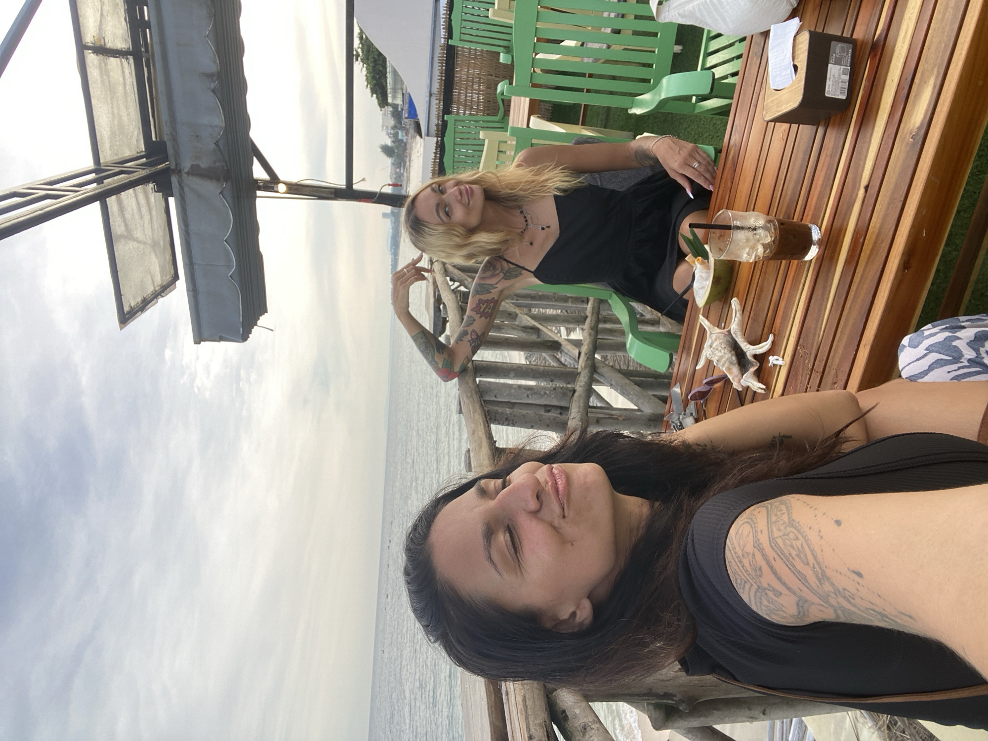 Two women are seated at a wooden table on a balcony overlooking the sea. The woman in the foreground is taking a selfie, her eyes closed and a slight smile on her face. She has dark hair and a tattoo on her arm. The woman in the background is wearing a black dress and has blonde hair. She is looking at the camera with her hand raised. On the table are two drinks, a bowl of fruit, a seashell, and a tissue box. The balcony is made of wood and has a railing. Beyond the balcony is a sandy beach and the ocean. In the distance, a city skyline can be seen. The sky is cloudy. The location is Phu Thuy Ward, Vietnam.