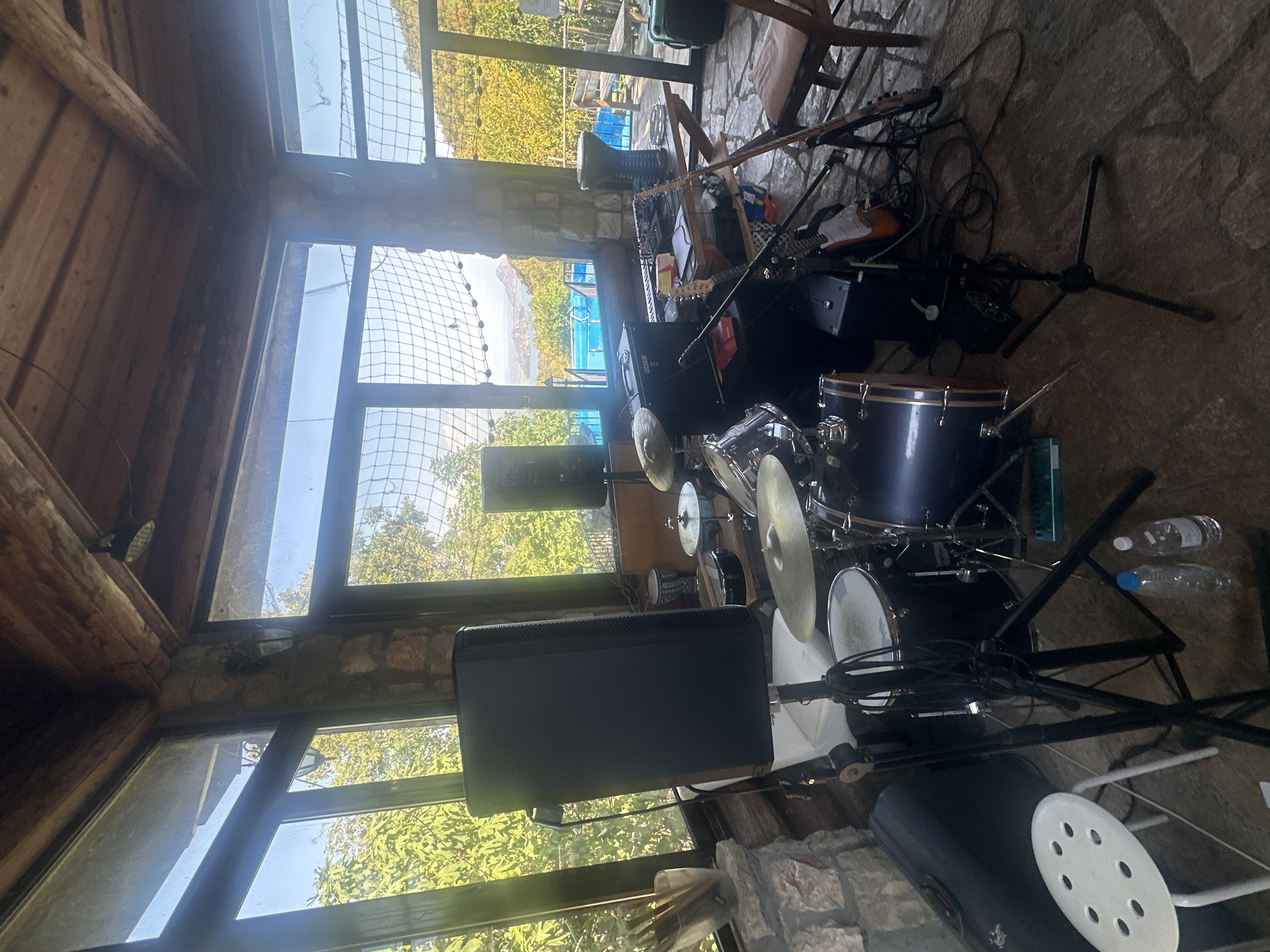 The image depicts an indoor music performance setup, likely within a structure with large windows offering a view of greenery and a distant body of water and hills, suggesting a scenic location, possibly on the outskirts of Cetinje, Montenegro. The room features a rustic aesthetic with exposed wooden beams and stone walls.
The central focus is a drum kit, complete with multiple drums, cymbals, and drumsticks held in a glass container on the left. Various audio equipment is scattered around: a large black speaker stands prominently on the left, another similar speaker is visible in the background, and a keyboard with a "BOSS" logo is on a table to the right. Two electric guitars are also present, one leaning against the table and another partially visible.
Other objects in the scene include a white stool in the foreground, two plastic water bottles, a dark carrying case, and a wooden chair. The floor appears to be made of stone. The overall impression is a ready-to-use stage or rehearsal space.