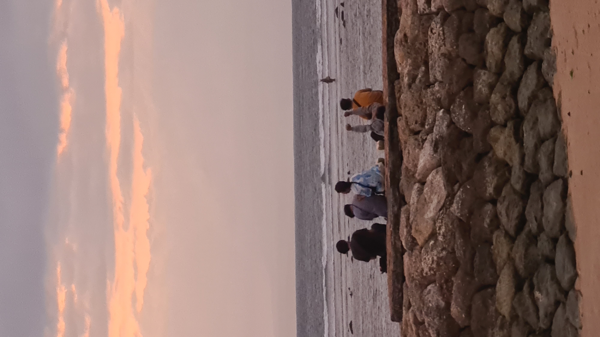 The image depicts a tranquil scene at a beach in Denpasar, Indonesia, likely during sunset or twilight, given the warm, diffused lighting and the soft colors of the sky. In the foreground, a rough-hewn stone embankment made of large, irregular rocks forms a barrier between the sandy beach and the ocean.
On top of this embankment, five individuals are seated, facing the water. From left to right, the first person is wearing a dark top. The second and third individuals are dressed in lighter, possibly striped or patterned clothing, with the third person wearing a blue and white patterned garment. The fourth and fifth individuals are positioned further to the right. The fourth person appears to be wearing a light-colored top, and the fifth person is wearing a bright yellow or orange shirt. Their postures suggest relaxation and observation of the sea.
The middle ground shows a sandy shore stretching towards the ocean. Further out, the water is a muted gray with gentle waves breaking, creating white lines of foam. In the distance, a lone figure is visible standing in the shallow water, adding a sense of scale and solitude to the expansive seascape.
The sky above is a soft canvas of pastel colors, with streaks of peach and pink from the setting sun or rising moon, interspersed with muted gray clouds. The overall atmosphere is serene and contemplative, capturing a moment of quiet enjoyment of the coastal environment in Denpasar.