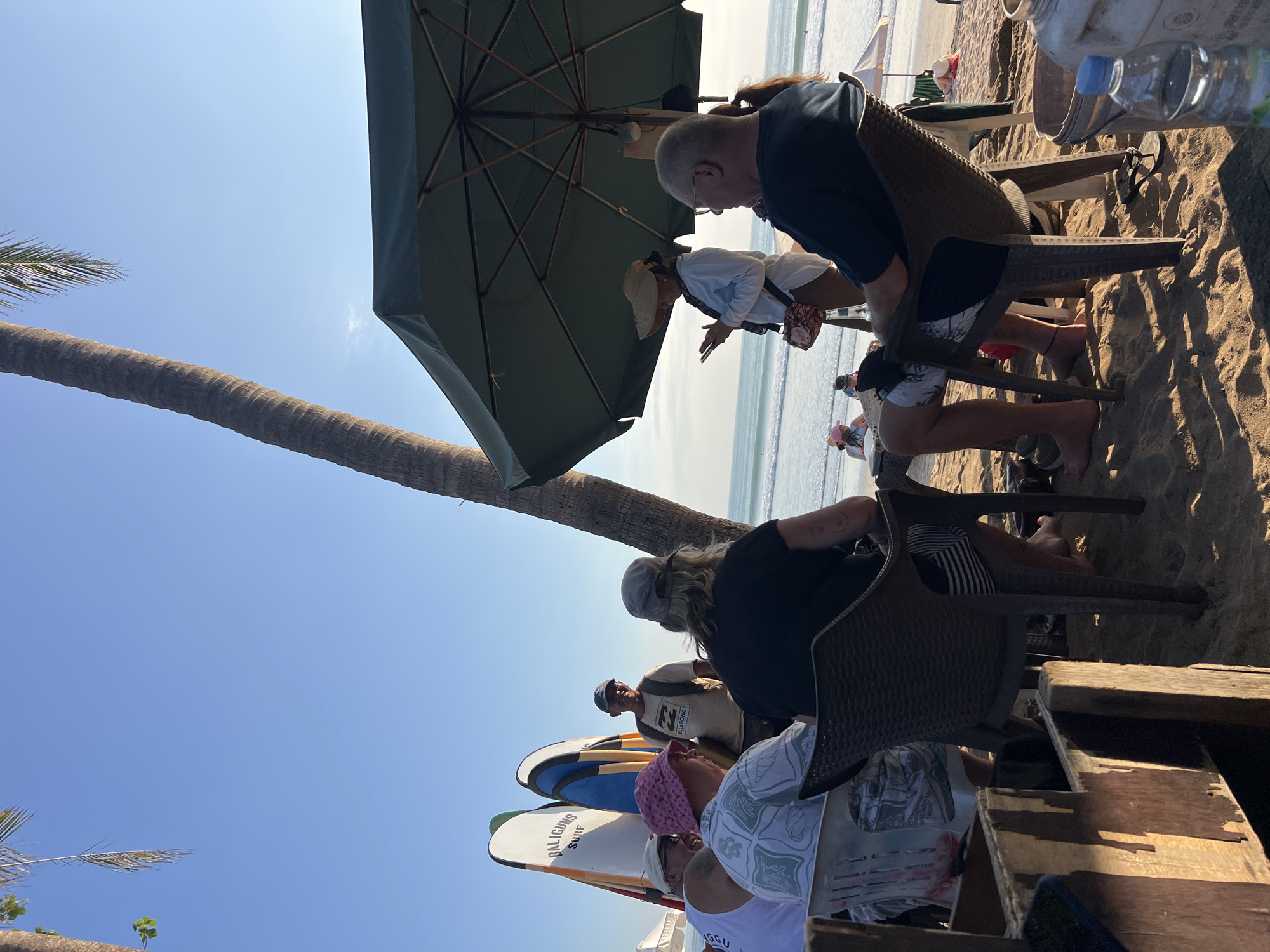 A group of people are gathered on the beach in Kuta, Indonesia, near the ocean. A tall palm tree is visible to the left, with a large, dark green umbrella providing shade over a table where several individuals are seated. Two surfboards stand upright near the group. The sand is a light brown, and the ocean waves are visible in the background. The sky is clear and blue. Some people are wearing casual clothing, and one person is holding a phone. The overall atmosphere appears relaxed, with people engaged in conversation or enjoying the beach setting.