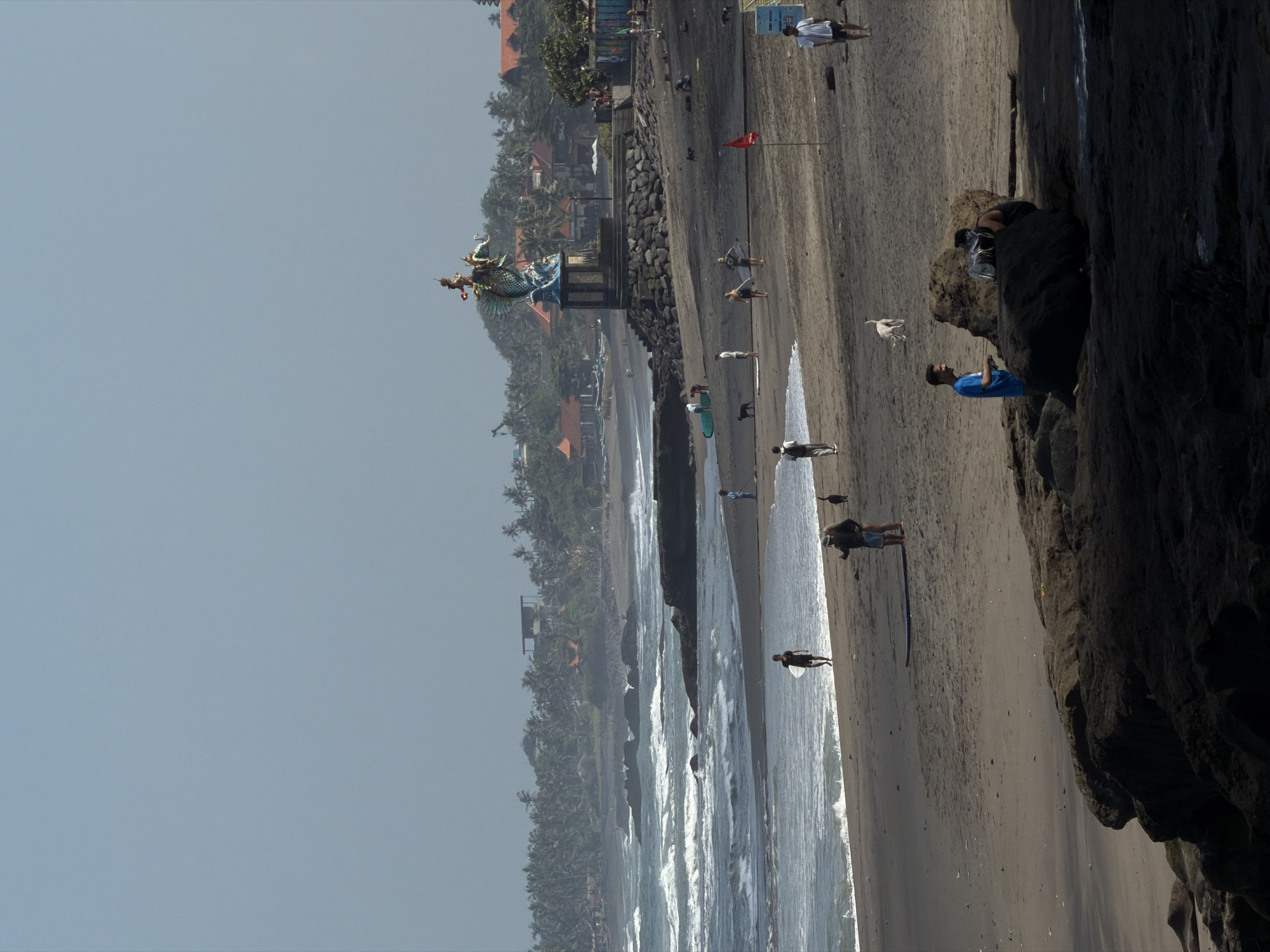 The image depicts a beach scene in North Kuta, Indonesia. The sky is hazy and light blue. In the background, a large, ornate statue stands on a raised platform overlooking the ocean, with lush green trees and buildings behind it. The beach itself is dark sand, with waves crashing on the shore. Several people are visible on the beach, some walking, some with surfboards, and some with dogs. A red flag is planted in the sand. The foreground is dominated by dark, rocky formations. The overall atmosphere suggests a sunny day with moderate activity on the beach.