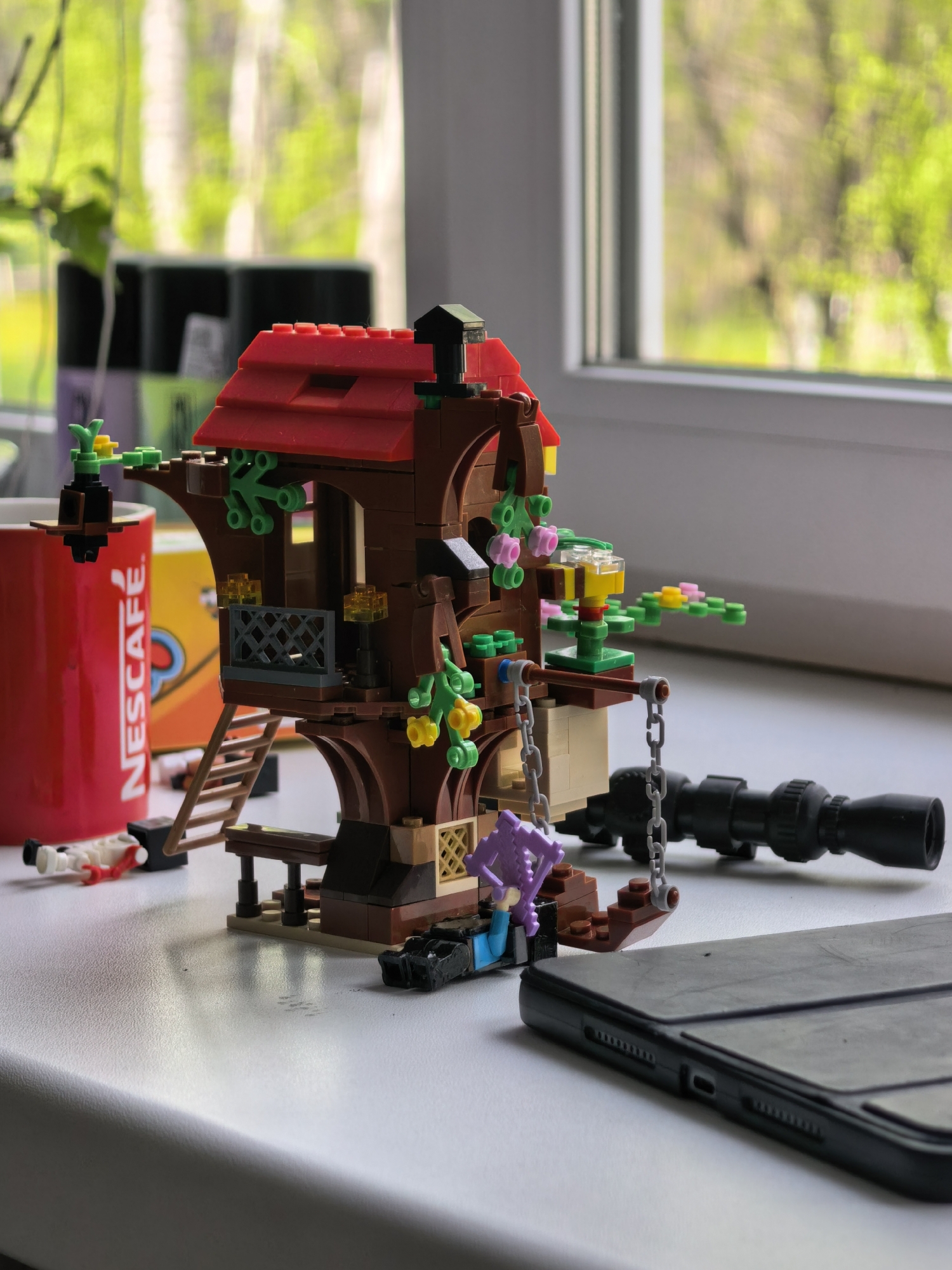 A Lego treehouse model stands on a white surface near a window with green foliage visible outside. A red Nescafe cup and a Nescafe box are partially visible behind the treehouse. A Lego minifigure lies on the surface near the cup, seemingly dismembered. A black telescope and a black smartphone are also placed on the white surface next to the treehouse. The scene is set indoors, possibly on a windowsill. The location of Sterlitamak, Russia is not evident in the image.