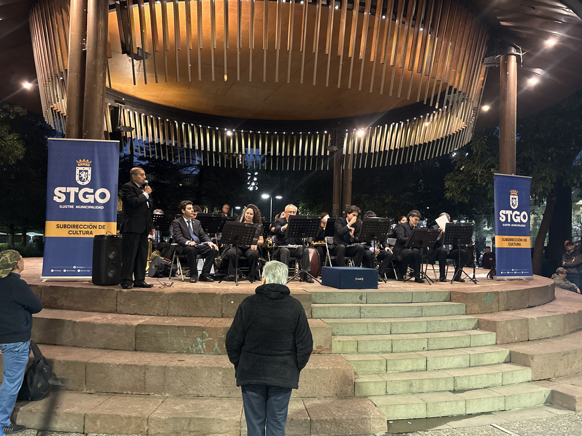 The image captures an outdoor concert taking place at night within the Parque Quinta Normal in Santiago, Chile. In the foreground, a person wearing a black jacket and blue jeans stands with their back to the camera, looking towards a band performing on a raised circular stage. The band, comprised of individuals in formal attire, is seated and playing various musical instruments. A man, identified by the "STGO ILUSTRE MUNICIPALIDAD SUBDIRECCIÓN DE CULTURA" banner to his left, stands center-stage addressing the audience with a microphone. The stage structure features a curved wooden roof illuminated by warm lights and vertical slats. The audience appears to be seated on tiered stone steps leading up to the stage. The overall scene is one of a community event, likely a cultural performance hosted by the municipality.