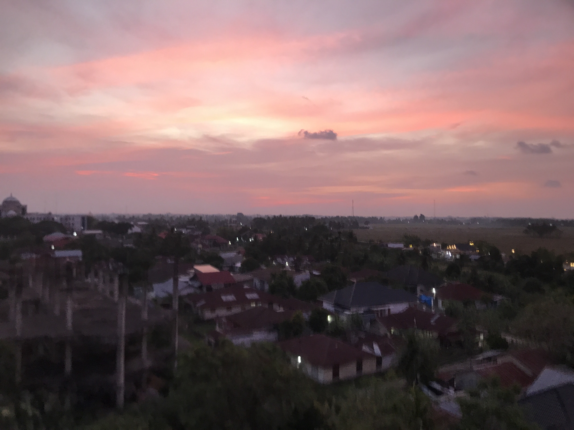 The image captures an expansive, elevated view of a residential and agricultural landscape in Cotbadatunong, Indonesia, bathed in the soft, vibrant light of either sunrise or sunset. The upper half of the frame is dominated by a dramatic sky, filled with diffused, streaky clouds painted in striking hues of pink, orange, and pale yellow. A more distinct, darker cloud formation is visible in the upper-middle right.
Below the colorful sky, the scene transitions to a sprawling settlement. Numerous houses with varying roof colors, primarily shades of red and brown, are nestled amidst abundant green trees and lush vegetation. Some tall, slender, palm-like trees are visible in the foreground, reaching towards the sky. Many houses exhibit small, bright artificial lights, suggesting the low ambient light of dusk or dawn. On the far left horizon, a large domed building, possibly a mosque, is faintly visible. Towards the right background, the landscape opens up into flat, expansive areas indicative of agricultural fields stretching into the distance.
No specific activity or event is taking place; the image presents a serene and tranquil snapshot of the area. The soft focus and warm, ambient lighting contribute to a peaceful atmosphere. There is no visible text within the image.