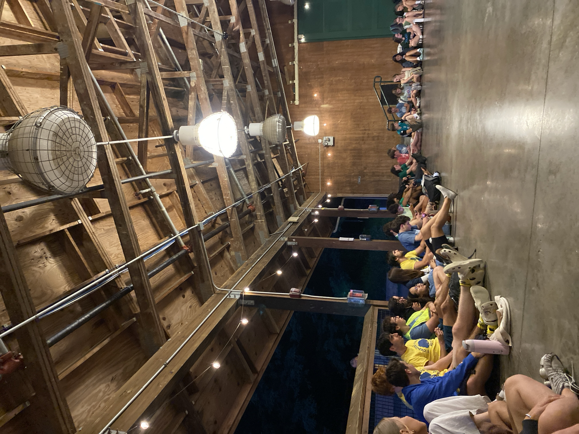 In Washington Township, United States of America, a group of young people are sitting on a concrete floor in a large, open-air pavilion with a wooden roof structure. The pavilion appears to be part of a camp or recreational facility, with exposed wooden beams and rafters overhead. Several large industrial-style light fixtures hang from the ceiling, along with string lights that provide ambient illumination. The group is seated in a semi-circle, facing inwards, with some individuals looking at a person or an area outside the frame of the image, while others are engaged in conversation. Most of the individuals are wearing casual clothing, including t-shirts, shorts, and athletic shoes. One person is holding a pink water bottle. The background of the pavilion is lined with wooden walls, and beyond that, a dark night sky with silhouettes of trees is visible. A set of stairs is located on the right side of the pavilion. The overall atmosphere suggests a relaxed gathering or event.