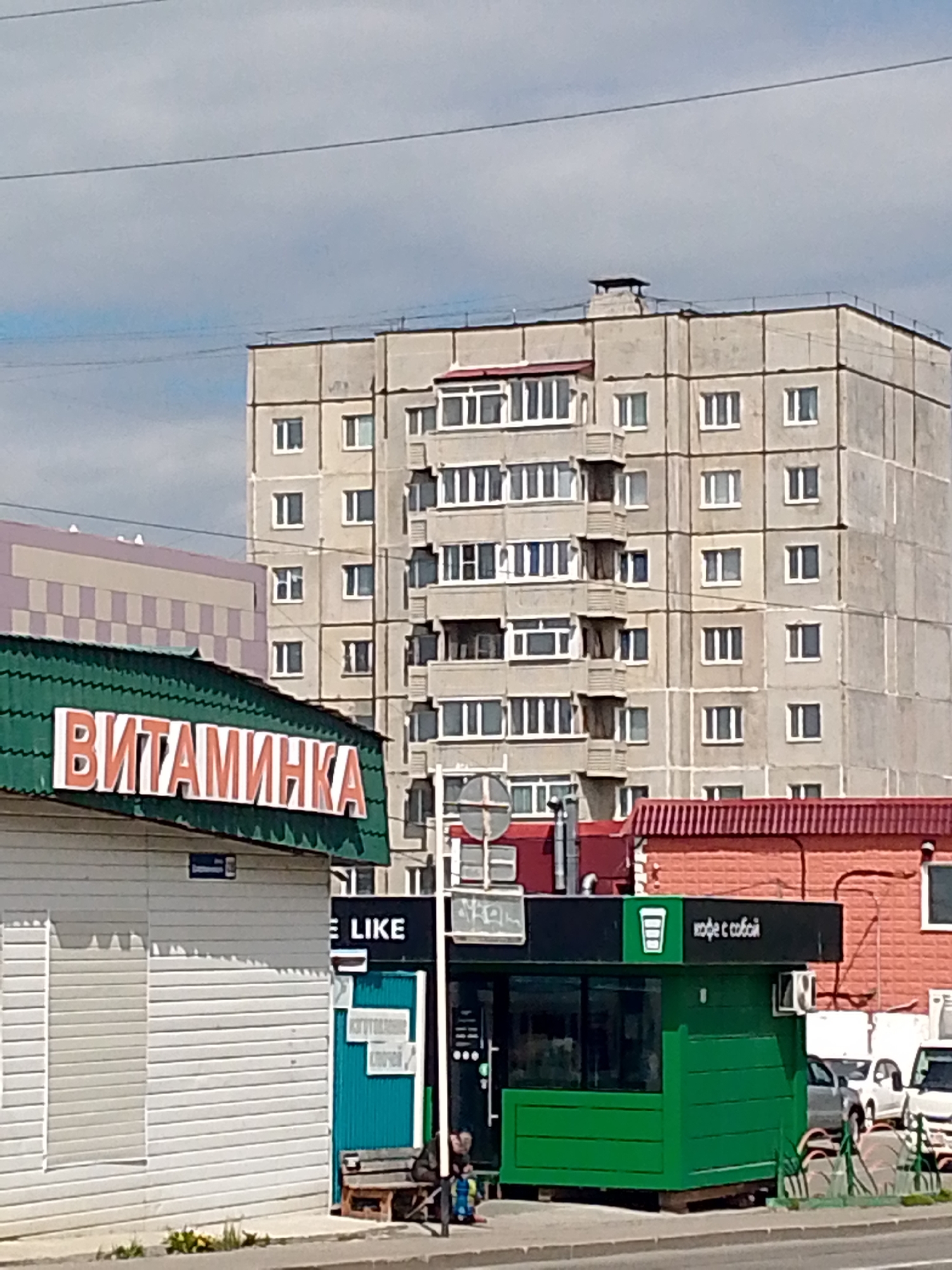 The image shows a street view in the city of Magadan, Russia. In the foreground, a green kiosk with the text "КОФЕ С СОБОЙ" (coffee to go) is visible, with cars parked behind it. To the left of the kiosk, a white building with a slatted facade is partially visible, and above it, a green awning with orange lettering reads "ВИТАМИНКА" (Vitaminka). Further in the background, a tall, grey apartment building with multiple windows dominates the skyline. The sky is overcast with scattered clouds. A bench is located next to the kiosk.