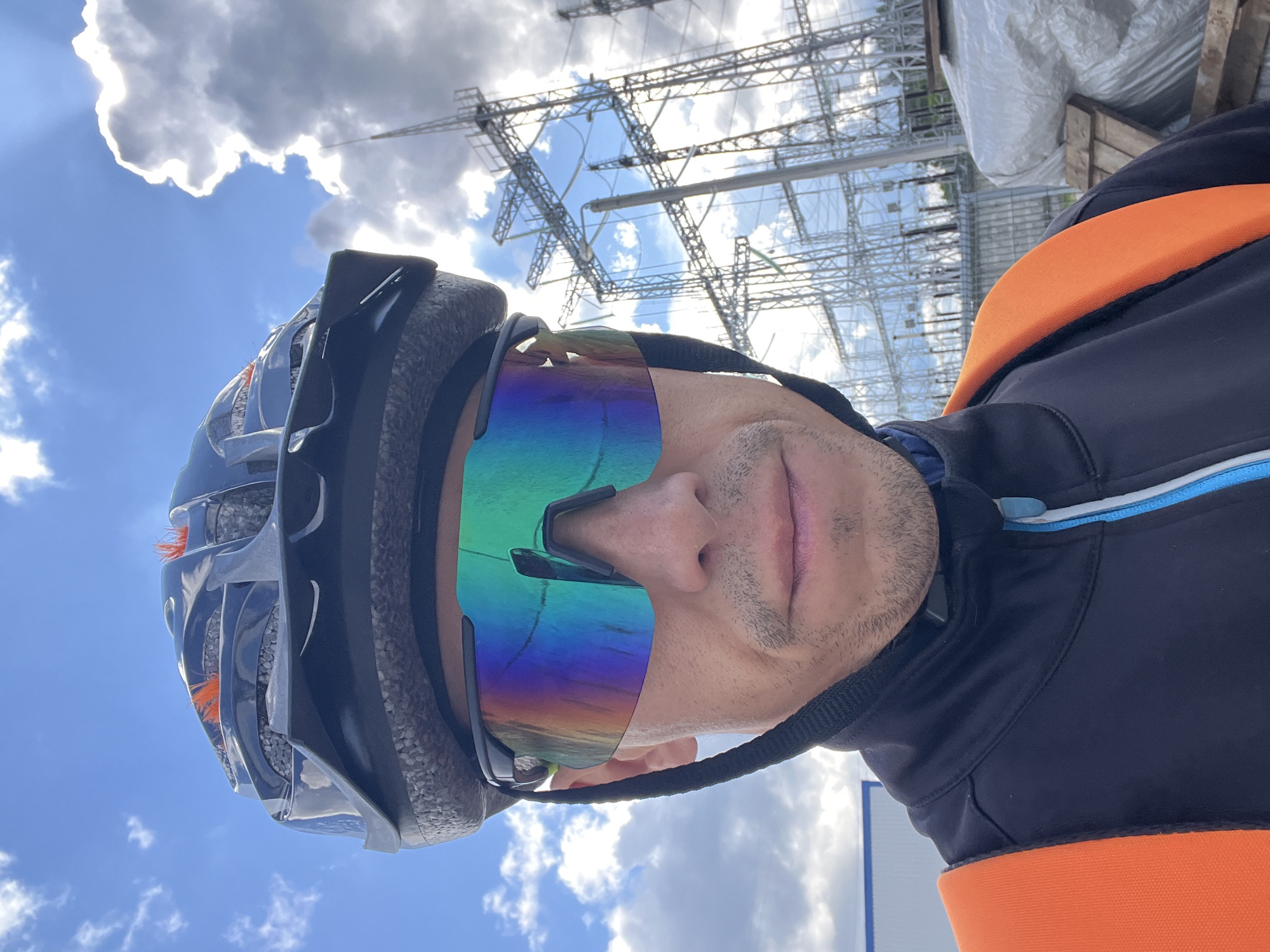 A close-up, slightly low-angle photograph captures a person wearing cycling gear against an outdoor background. The individual, seen from the chest up, has their face mostly obscured by large, highly reflective, iridescent sunglasses that display a rainbow spectrum of colors. Stubble is visible on their chin and upper lip. They are wearing a dark grey or black cycling helmet with orange internal padding visible through its vents. Their attire includes a dark blue or black long-sleeved jersey or jacket featuring a distinct horizontal orange band across the upper back/collar area, with subtle light blue and white piping accents.
The background consists of a bright blue sky with scattered, fluffy white and grey clouds. Sunlight streams through gaps in the clouds, creating visible rays. Dominating the upper and central background are extensive, intricate metal lattice structures, strongly resembling an electrical substation or high-voltage power transmission towers. Some white tarpaulins or covered objects are also visible on the right side of the frame. The scene is set outdoors on a sunny, partially cloudy day, appearing to be near industrial infrastructure in Chaikovsky, Russia. No discernible text is visible in the image.