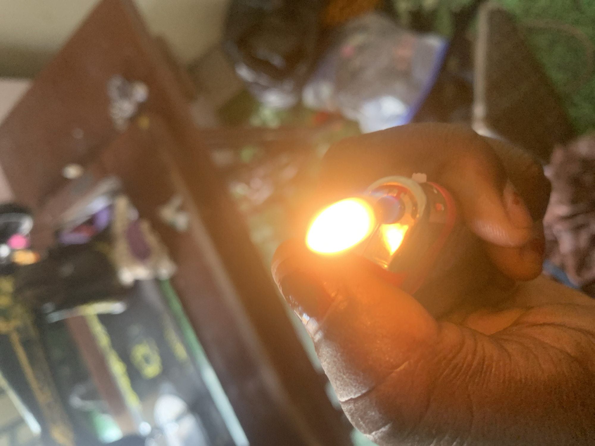 A hand holds a lit lighter, illuminating the foreground. In the blurred background, a sewing machine sits on a wooden surface, with various objects scattered around it, including bags and fabrics. The scene appears to be an indoor setting in Jauro Yinu, Nigeria.