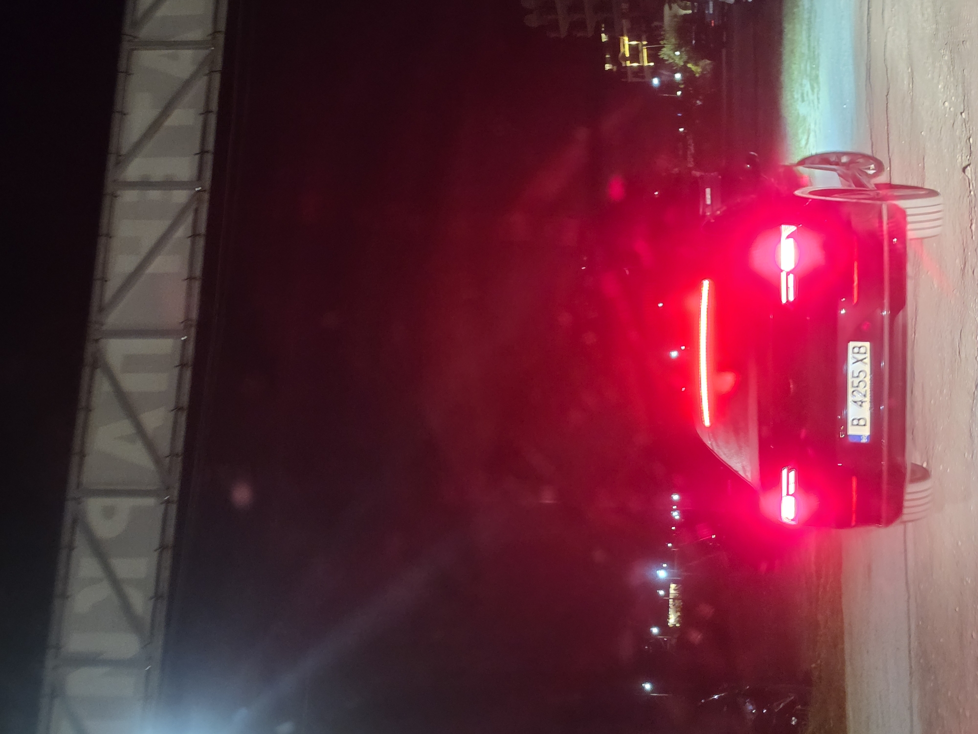 The image depicts a street scene at night in Kranevo, Bulgaria. In the foreground, a black car is stationary, its red taillights illuminated. The license plate of the car is visible and reads "B 4255 XB". The car is positioned on a paved surface, likely a road or parking area. In the background, there are illuminated streetlights and the indistinct outline of buildings. Above the scene, a large sign structure with metal bracing is visible against the dark sky. The overall lighting is low, with the red glow from the car's taillights dominating the foreground.