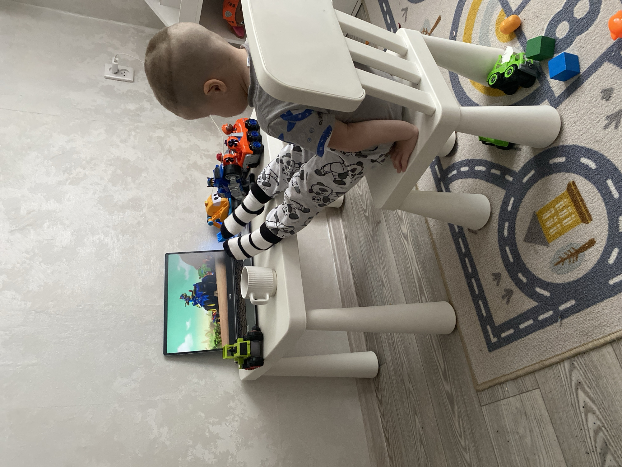 The media file captures a young child in an indoor setting, likely in Sochi, Russia. The child, with short light hair, is viewed from the back and side, sitting in a white high chair. They are wearing grey pajamas adorned with panda bear prints and black and white striped socks.
The child is intently focused on a laptop placed on a small white children's table directly in front of them. The laptop screen displays a colorful animated scene, possibly a cartoon featuring vehicles and characters. The brand "TECLAST" is visible on the bottom bezel of the laptop. A white ribbed ceramic mug rests on the table next to the laptop.
Around the child are various toys, including several toy vehicles resembling trucks and transforming robot figures. Some toys are lined up next to the high chair, while others, like a green toy truck and blue and orange building blocks, are scattered on the floor rug.
The floor beneath and around the high chair is a mix of light-colored plank flooring and a patterned children's rug featuring road and building designs. The wall in the background is light-colored with a textured, mottled finish. A white electrical socket with a charger plugged in is visible on the wall. The scene suggests a daytime setting indoors, with the child engaged in screen time, surrounded by their playthings in a domestic environment.