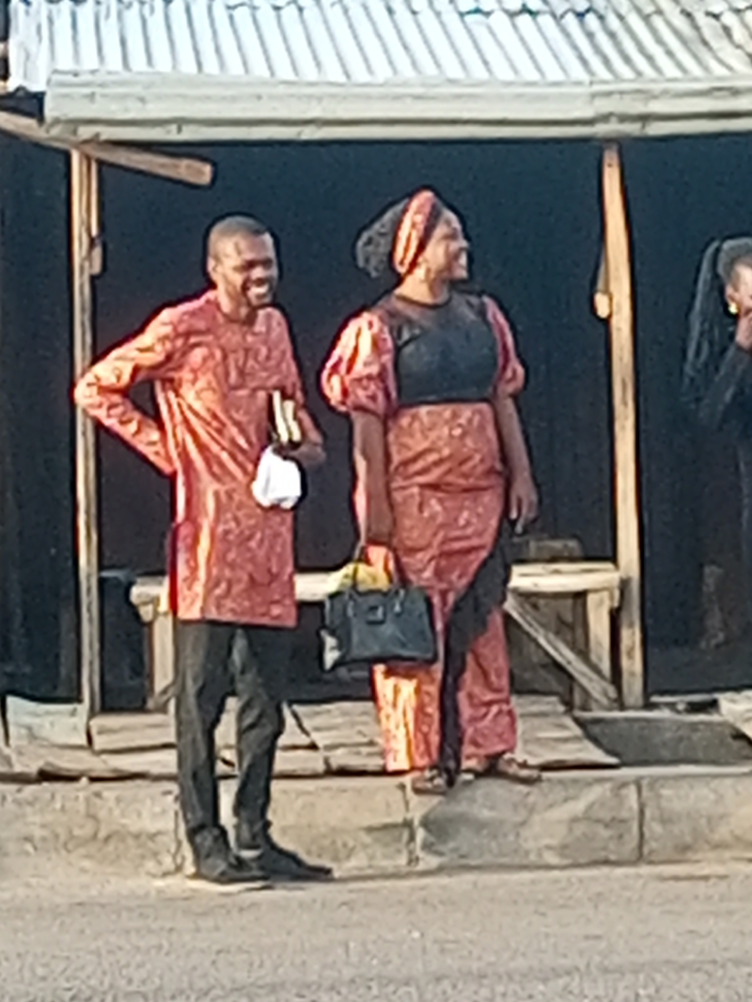The image depicts a street scene in what appears to be a West African urban area. A man and a woman are standing on a concrete curb in front of what looks like a small shop or stall with a tin roof. The man is wearing a patterned orange shirt and black pants. He has short hair and is smiling. He is holding something in his hand, possibly a small book and a white cloth. The woman is wearing a matching patterned orange outfit with a black top and head wrap. She is carrying a dark colored handbag. Both are looking off to the right. Another woman in black can be seen standing partially obscured behind the woman. The background of the stall is dark, and there's a rough wooden bench visible. The photo is slightly blurry. There is no direct confirmation that this is in Tempure, Nigeria, but the style of dress and general environment are consistent with that region.